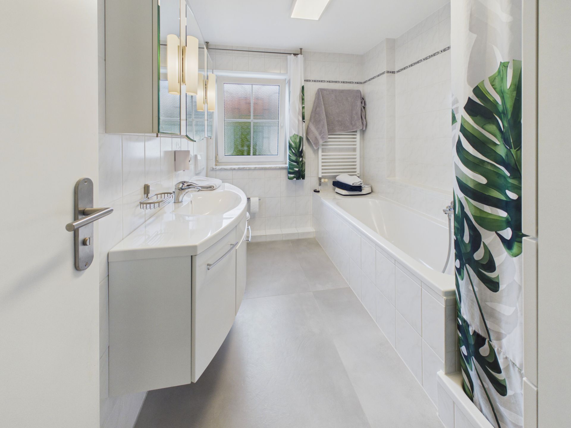 Helles Badezimmer mit Badewanne, Waschbecken, Spiegel und Fenster, Duschvorhang mit Blattmuster.