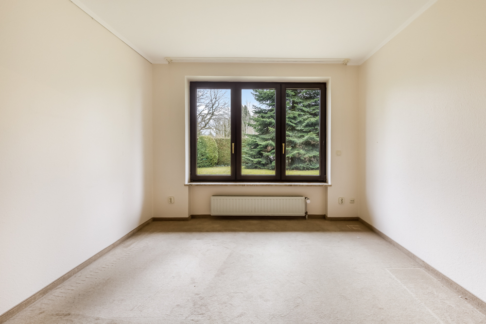 Leeres Zimmer mit großem Fenster, beige Wänden und Blick auf Garten mit Bäumen.