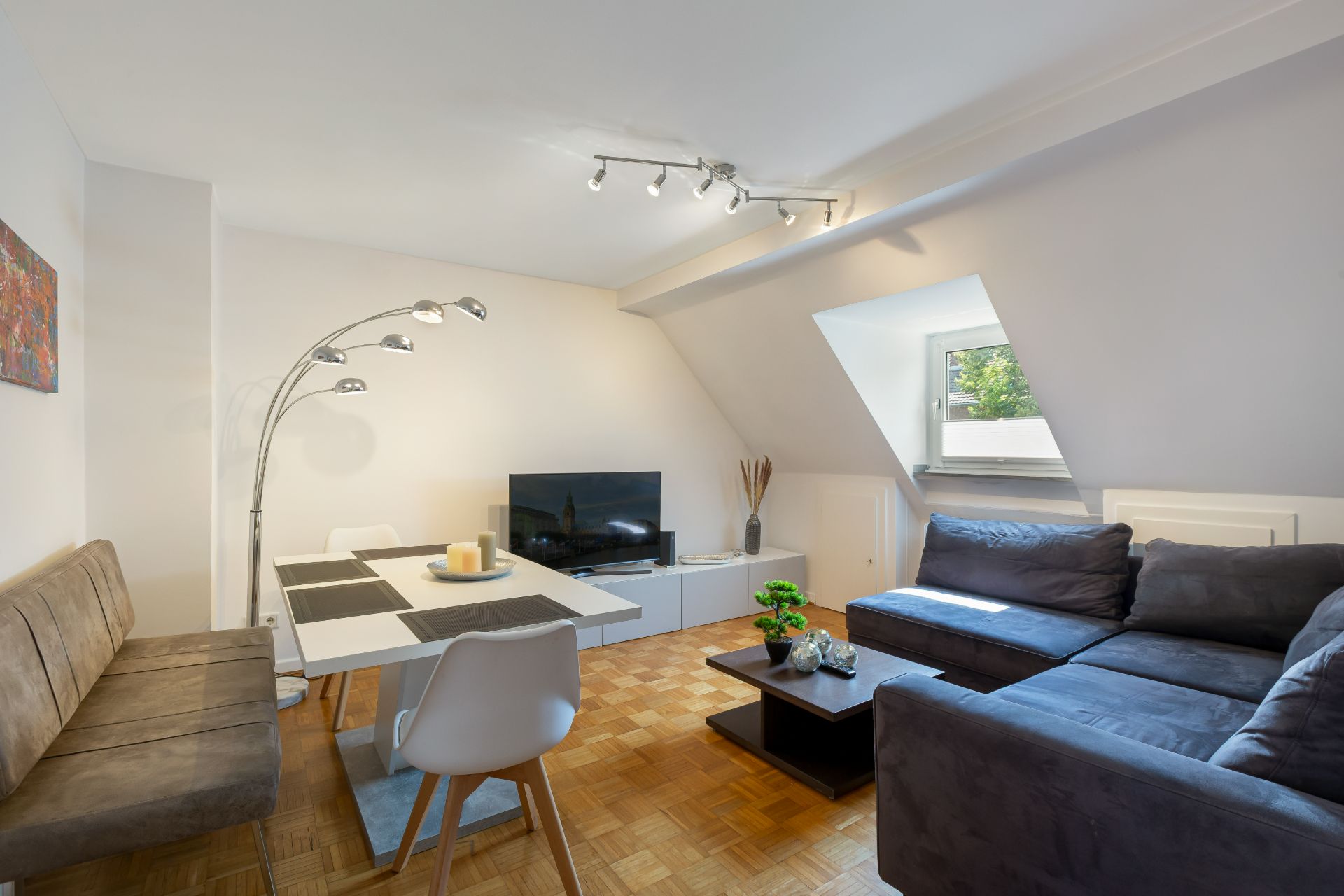 Wohnzimmer mit grauem Ecksofa, Esstisch, Fernseher und Fenster unter einer Dachschräge.
