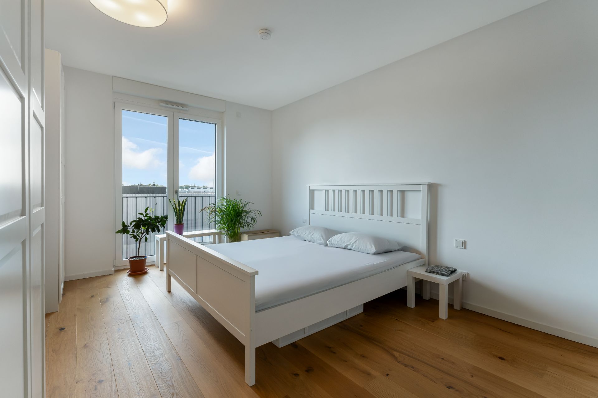 Helles Schlafzimmer mit Doppelbett, Holzboden, großen Fenstern und mehreren Zimmerpflanzen.
