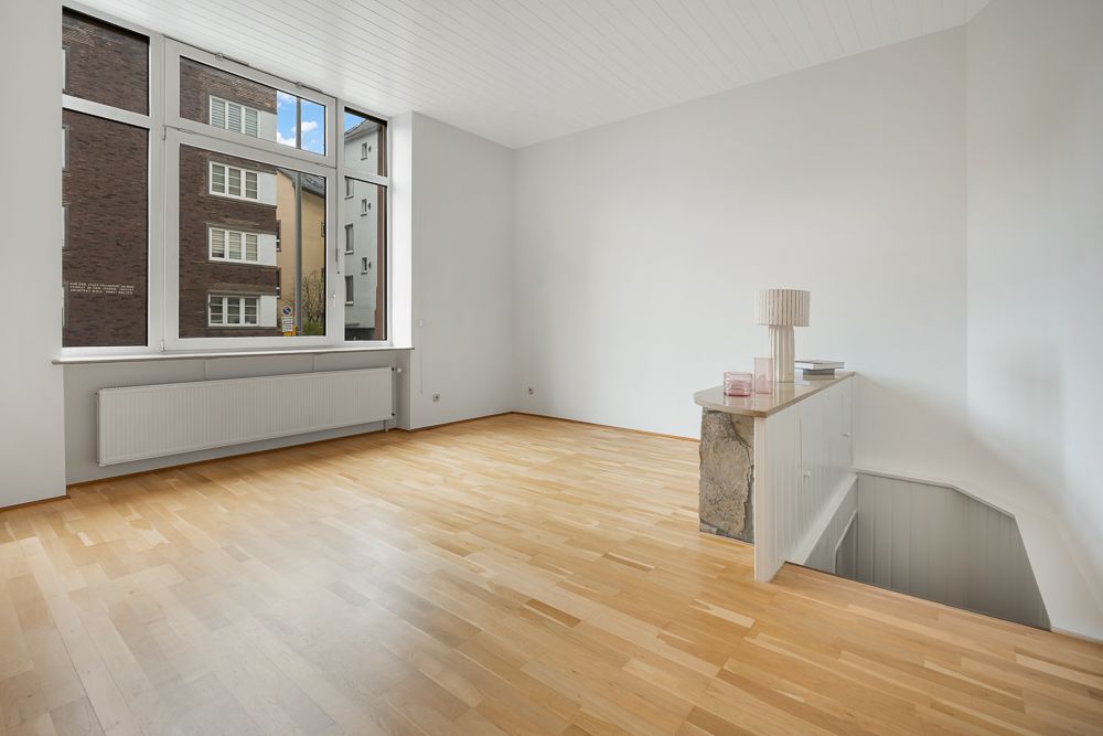 Helles Zimmer mit großem Fenster, Holzfußboden und Treppenabgang, minimal möbliert.