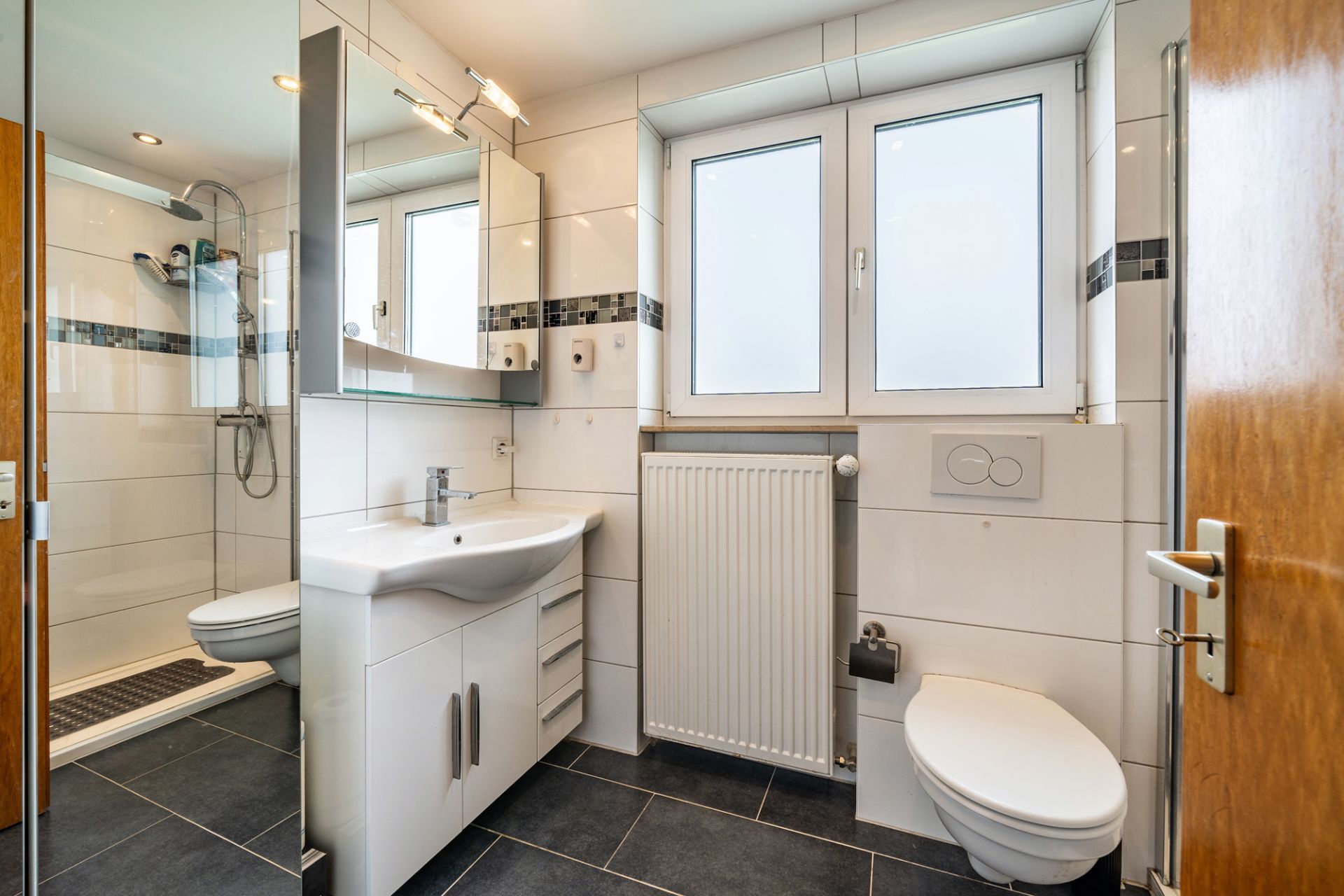 Modernes Badezimmer mit Waschbecken, Spiegel, Dusche, Toilette und Fenster.