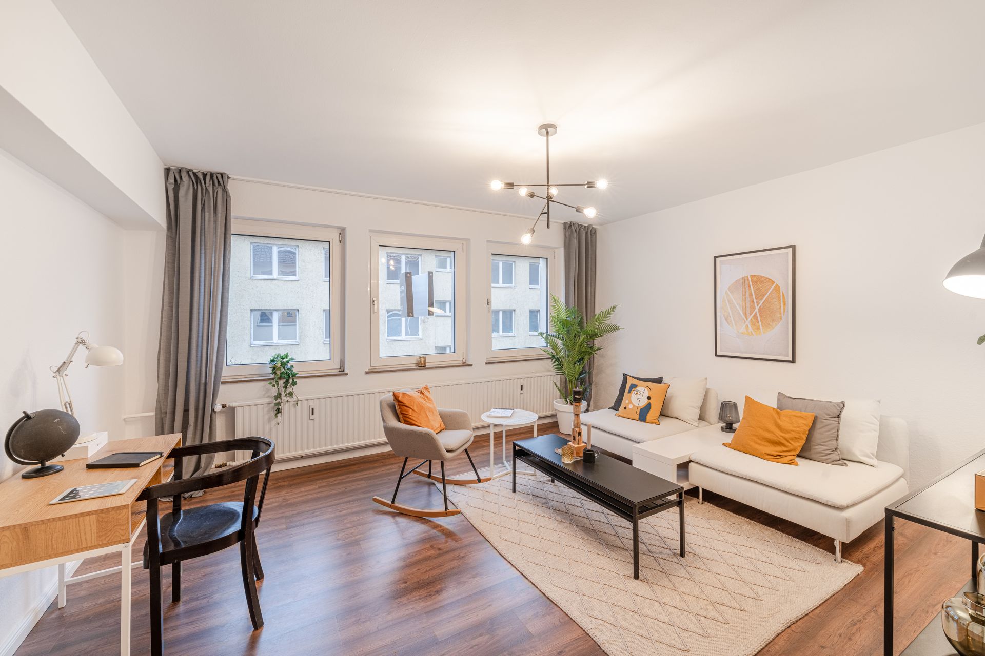 Helles Wohnzimmer mit Sofa, Schreibtisch, Sessel, Couchtisch und drei Fenstern.