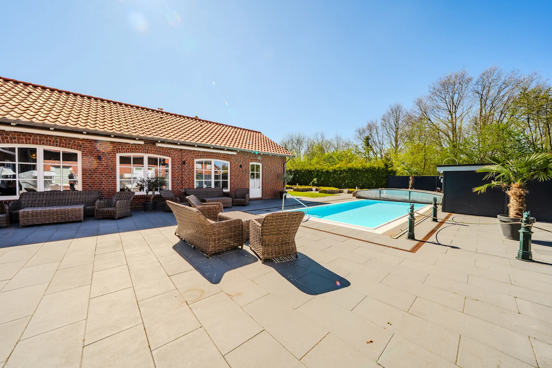 Große Terrasse mit Gartenmöbeln und Pool vor einem Backsteinhaus bei sonnigem Wetter.