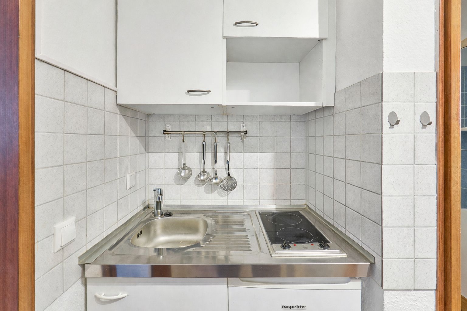 Kleine Küche mit Spüle, Kochfeld, Hängeschrank und Küchenutensilien an der Wand.