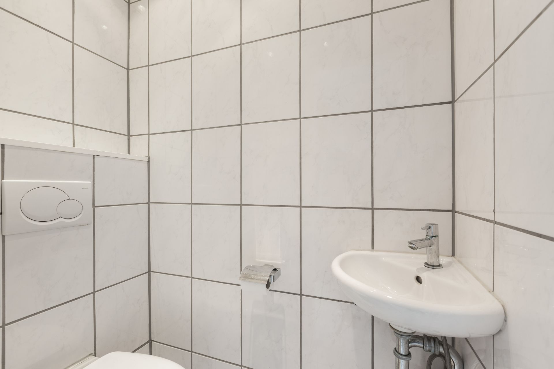 Kleines Badezimmer mit weißen Fliesen, WC, Eckwaschbecken und Toilettenpapierhalter.