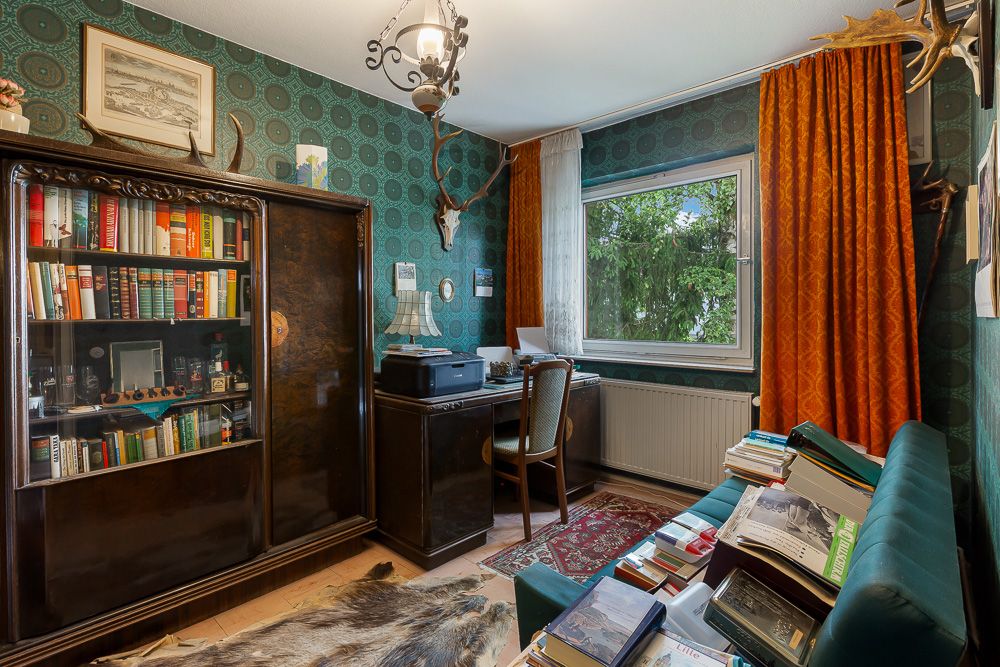 Kleines Arbeitszimmer mit Bücherregal, Schreibtisch, Sessel, Sofa, vielen Büchern und orangefarbenen Vorhängen.