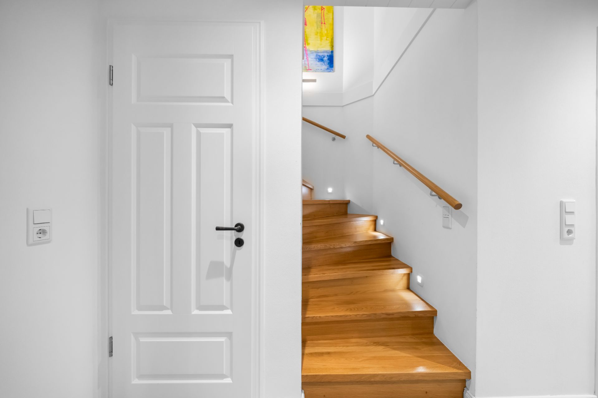 Holztreppe mit Handlauf neben geschlossener weißer Tür, Wand in Weiß gestrichen.