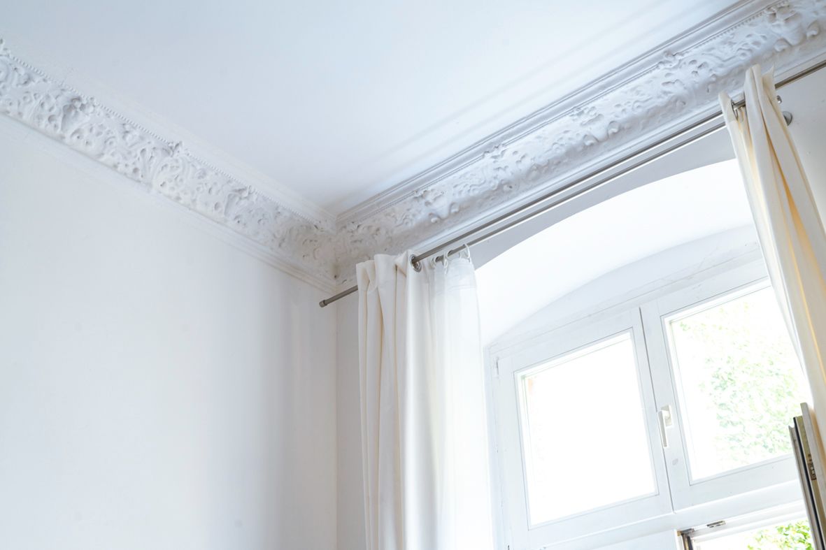 Weißes Zimmer mit Stuckdecke, Vorhangstange sowie Fenster mit weißen Gardinen.