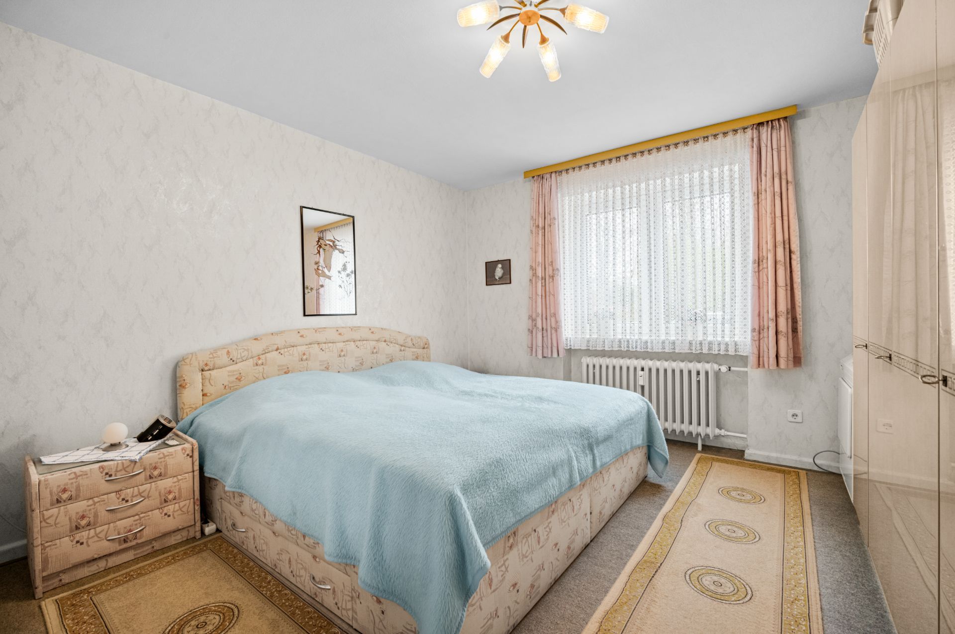 Schlafzimmer mit Doppelbett, Nachttisch, Fenster mit Vorhängen und Teppichen auf dem Boden.