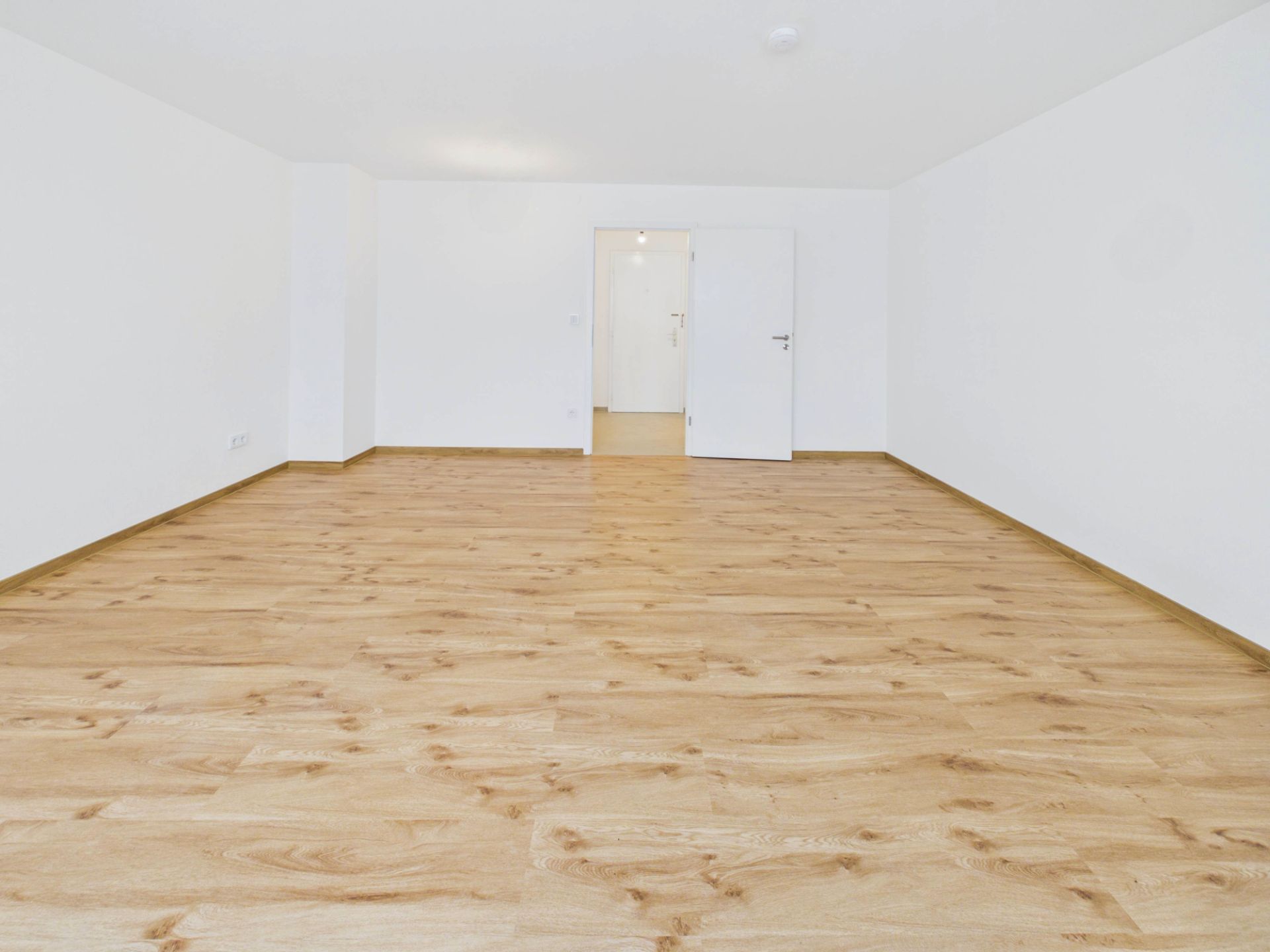 Leerer Raum mit hellem Holzfußboden und weißen Wänden, offene Tür im Hintergrund.
