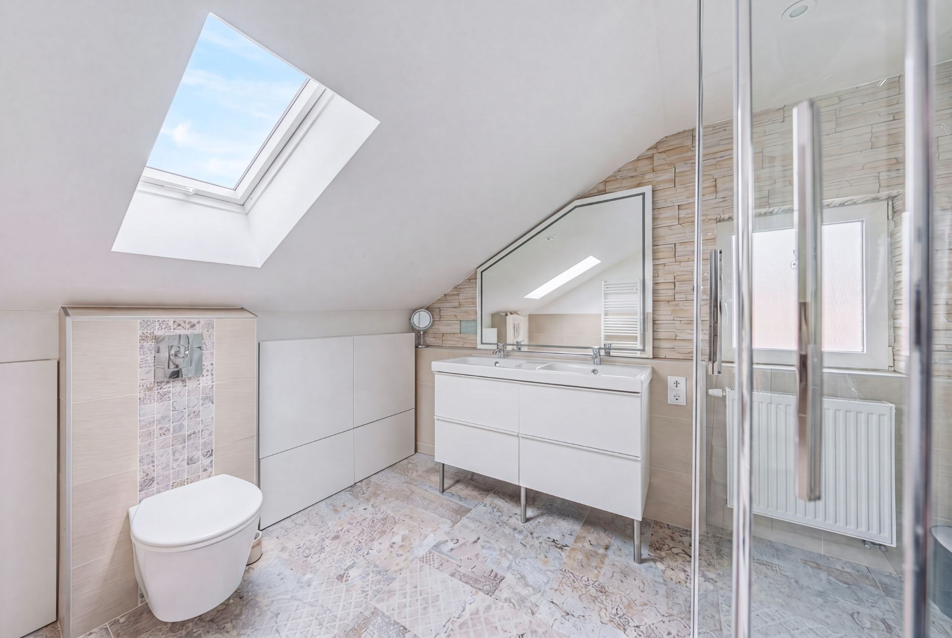 Helles Badezimmer mit Dachfenster, Waschbecken, Spiegel und wandhängender Toilette.