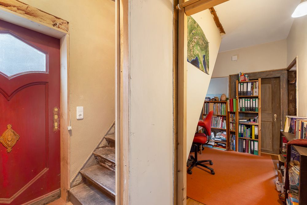 Flur mit roter Tür, Holztreppe und Blick in ein Arbeitszimmer mit Regalen und Schreibtisch.