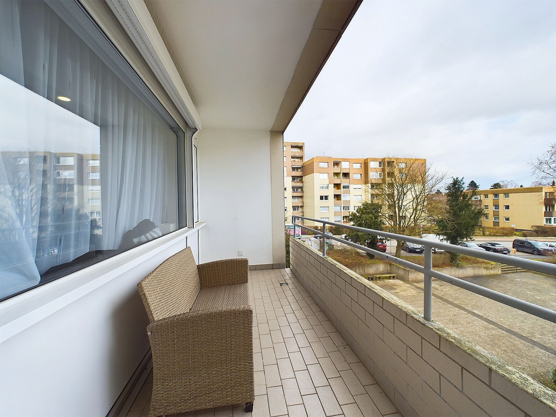 Balkon mit Rattansofa, Blick auf Wohngebäude und Parkplatz, große Fenster mit Vorhängen.