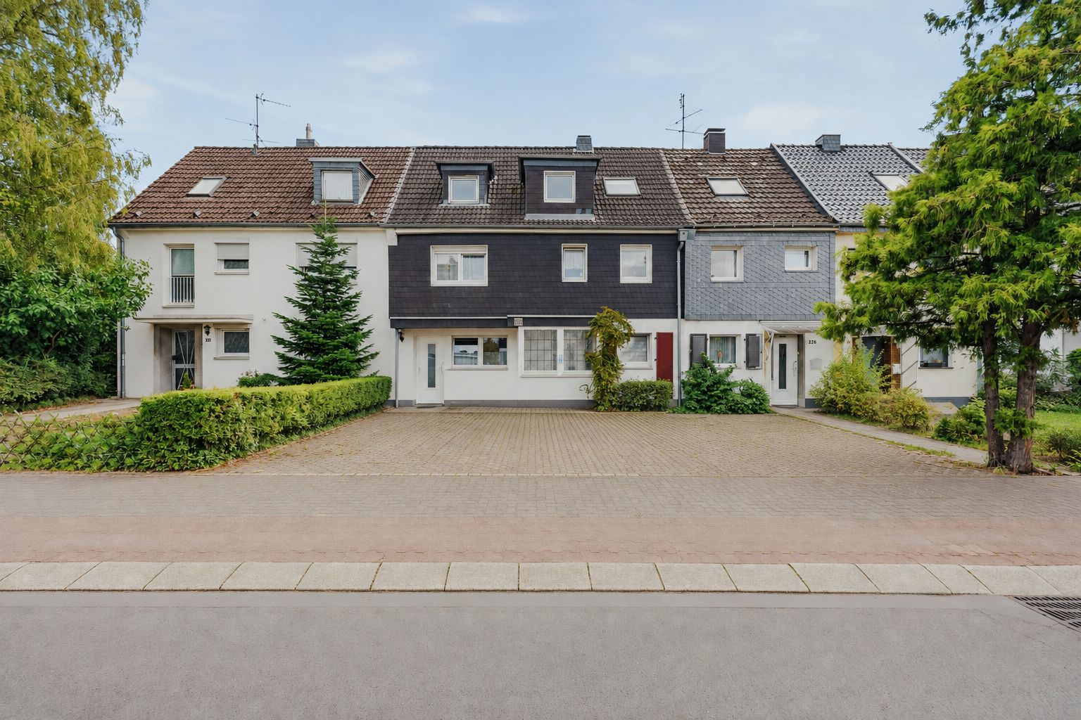 Mehrere Reihenhäuser mit Vorgarten und gepflastertem Parkplatz, Bäume und Büsche davor.
