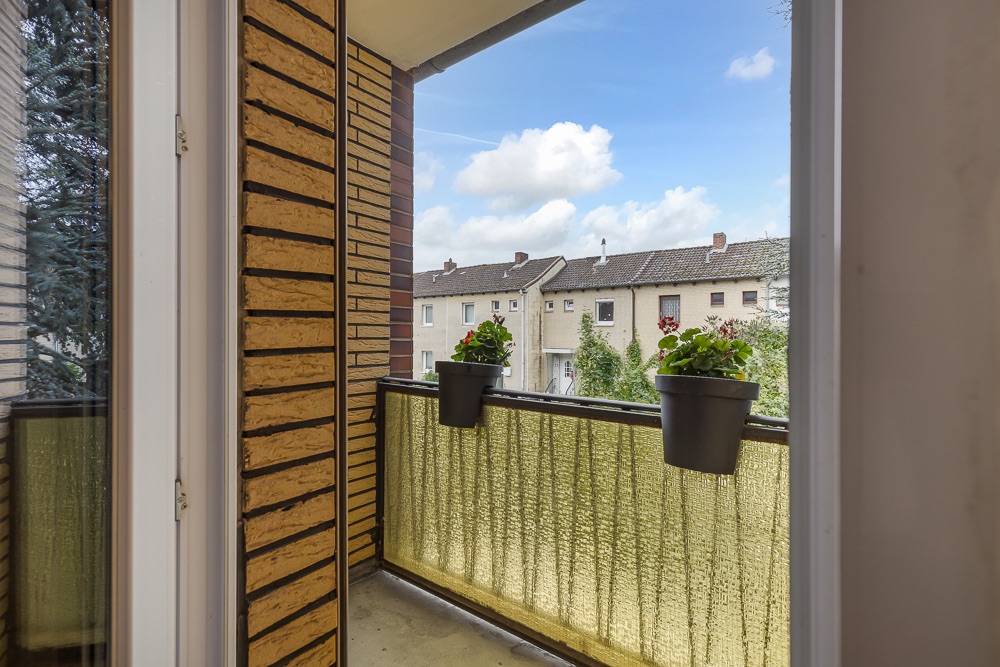 Kleiner Balkon mit zwei Blumentöpfen am Geländer, Ausblick auf Nachbarhäuser und blauen Himmel.