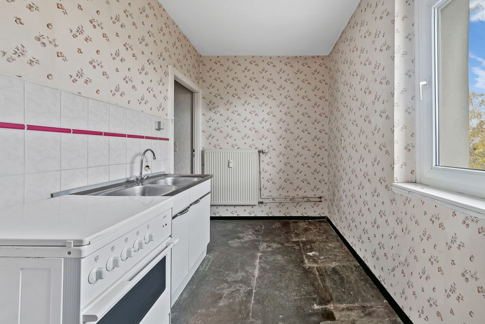 Leere Küche mit Herd, Spüle, floral gemusterten Tapeten und großem Fenster.