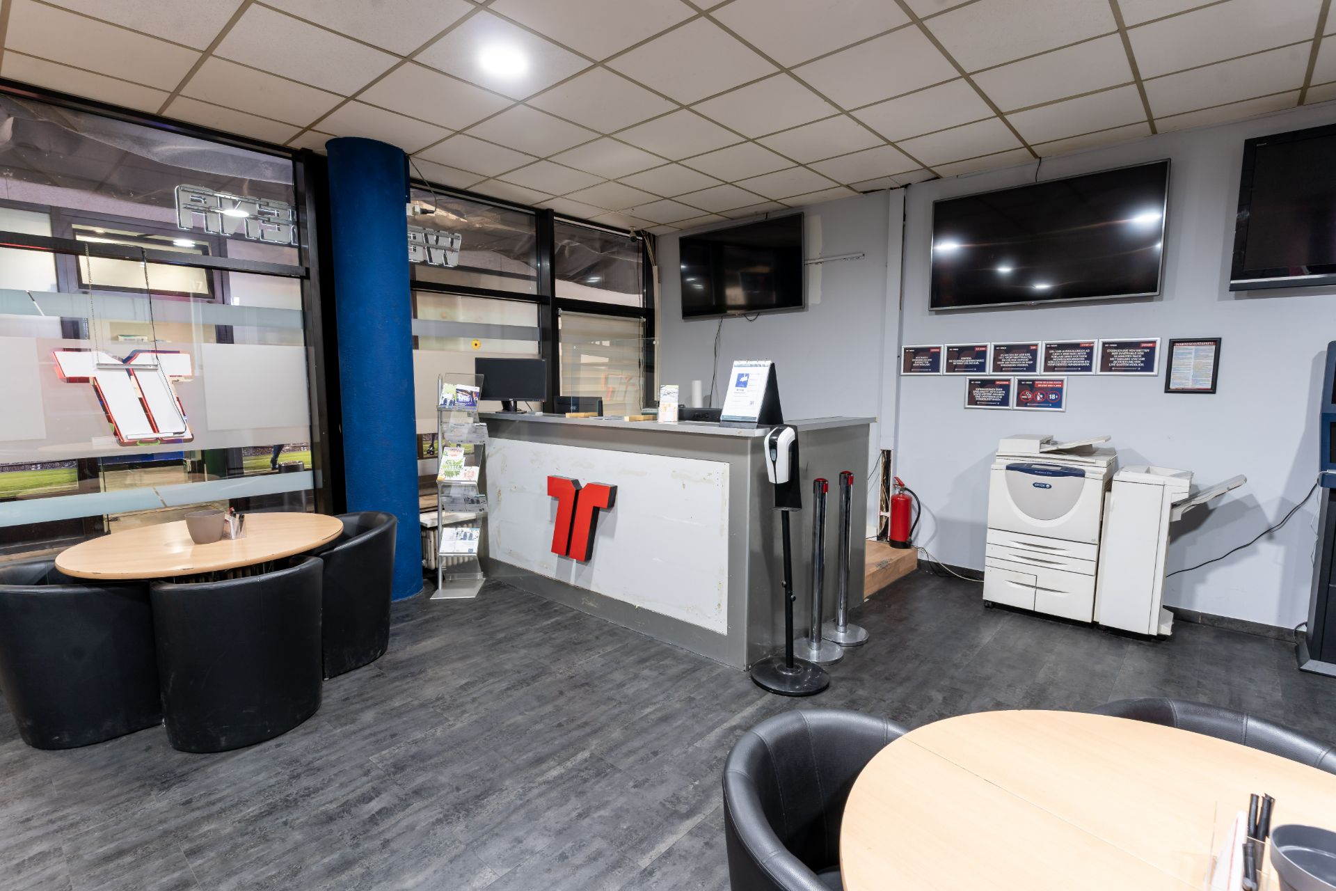 Empfangsbereich mit Tresen, Sitzgruppen, Fernsehern an der Wand und einem Drucker im Hintergrund.