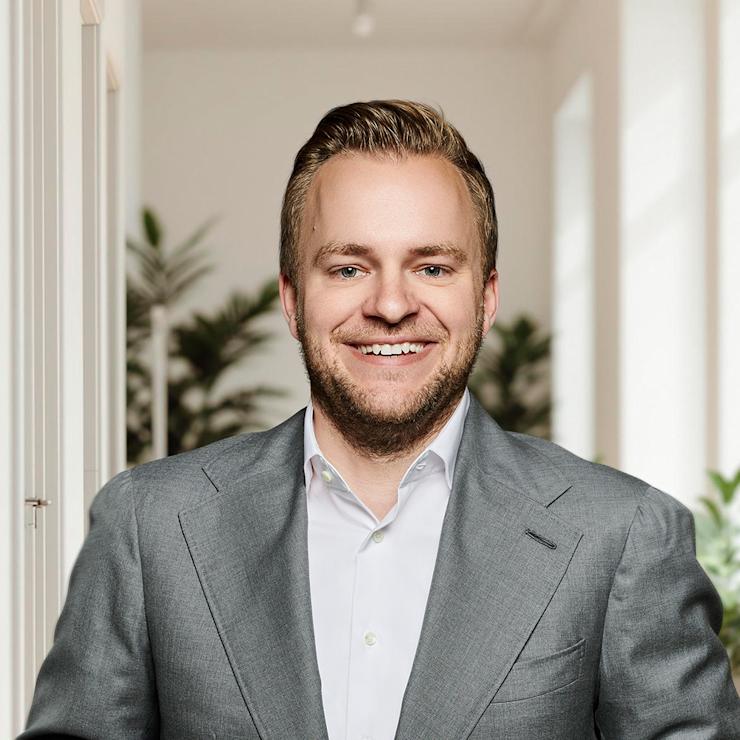 Mann im grauen Anzug steht in einem hellen Raum mit Pflanzen im Hintergrund.