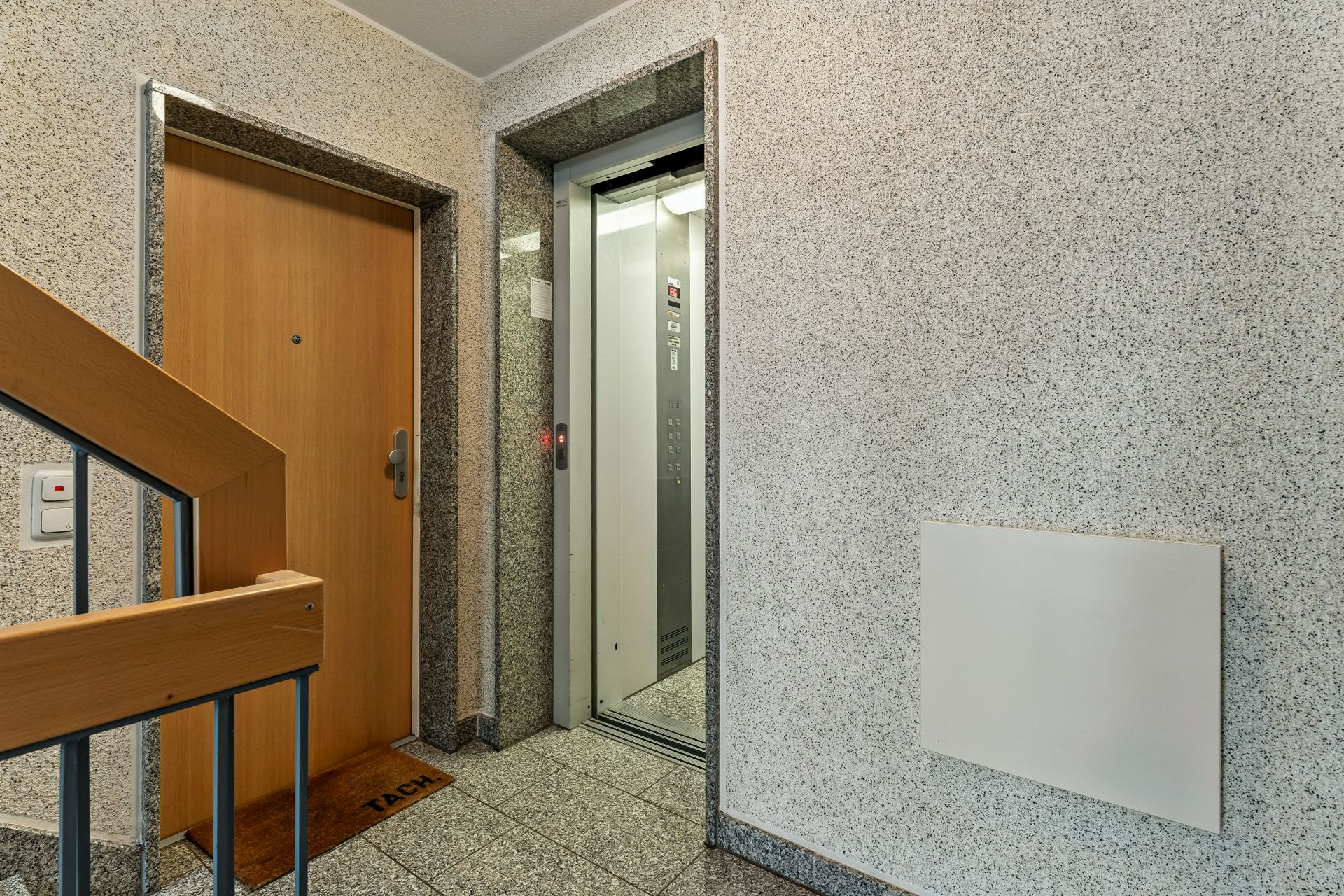 Treppenhaus mit Holztür links und offenem Aufzug rechts.