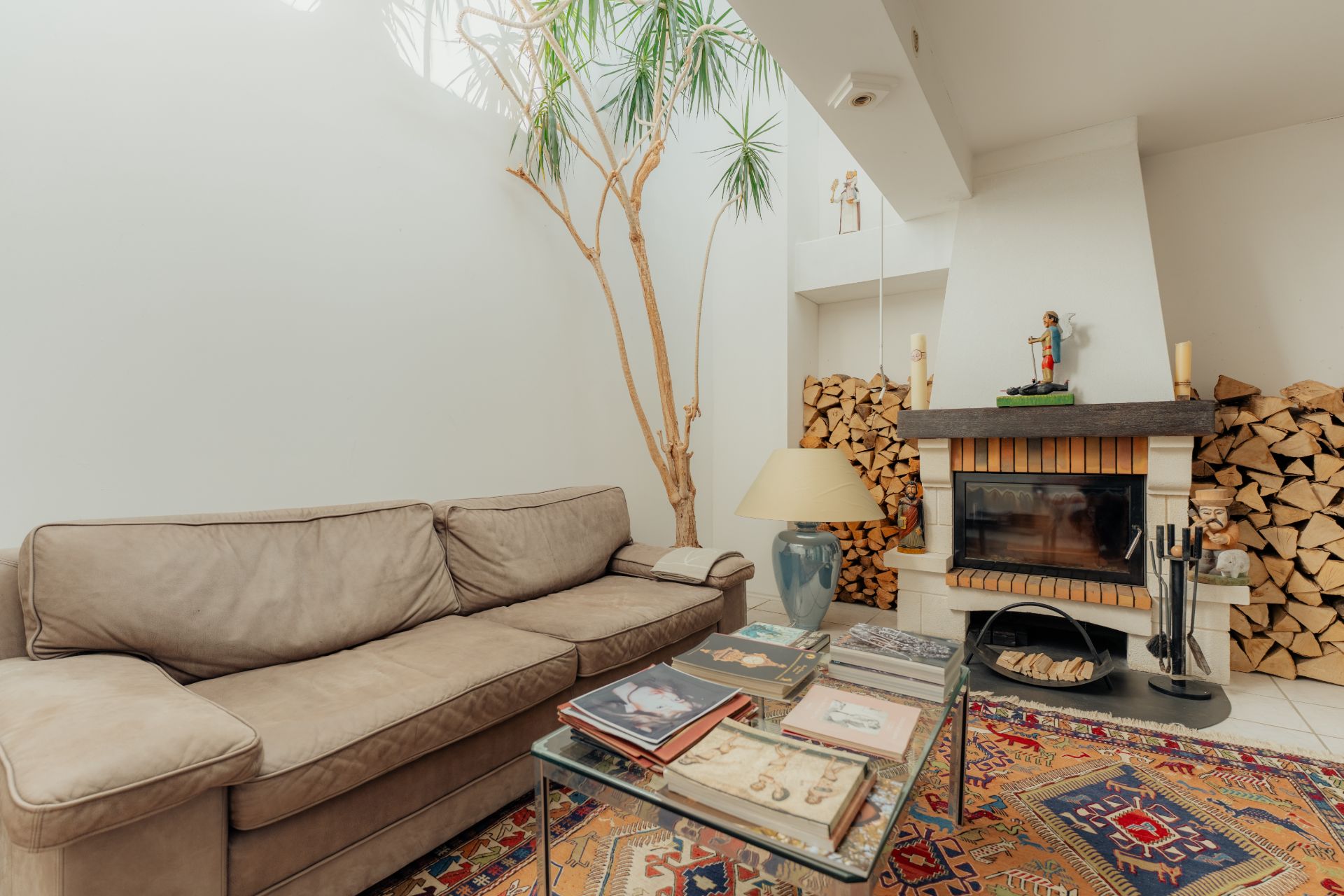 Wohnzimmer mit beigem Sofa, Glastisch, Kamin und gestapeltem Brennholz an den Wänden.