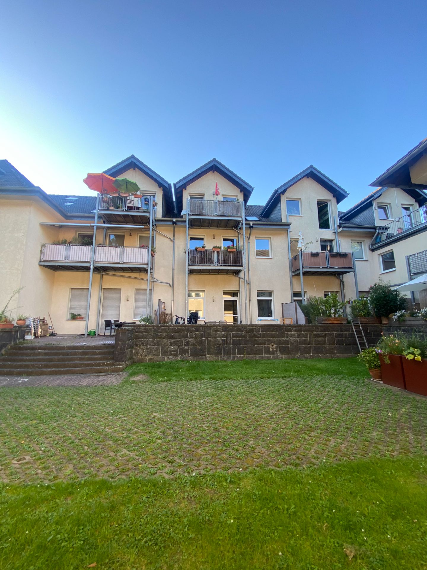Mehrfamilienhaus mit Balkonen und Gartenfläche im Vordergrund bei blauem Himmel.