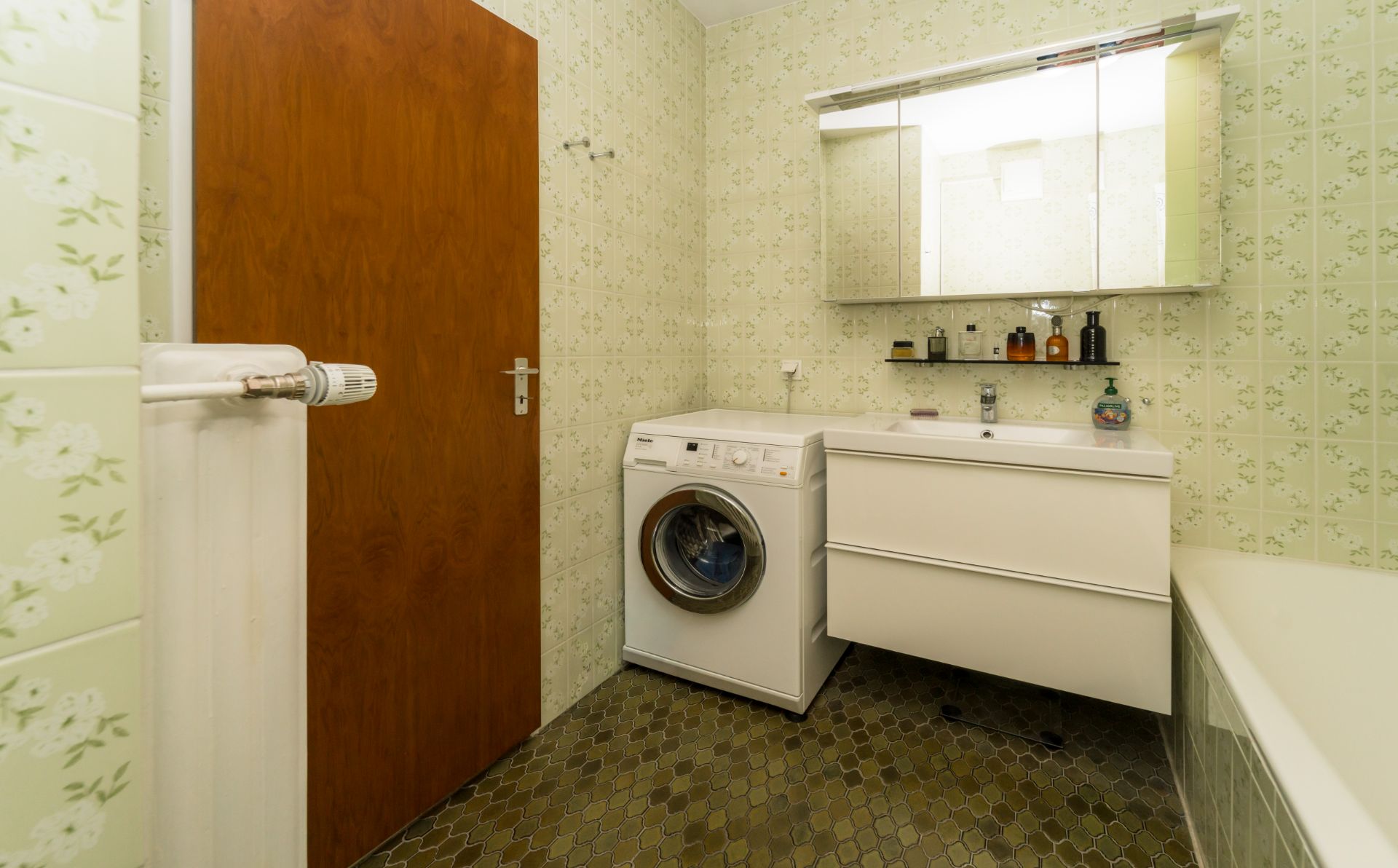 Badezimmer mit Waschmaschine, weißem Waschbecken, Spiegel und grünen Fliesen.