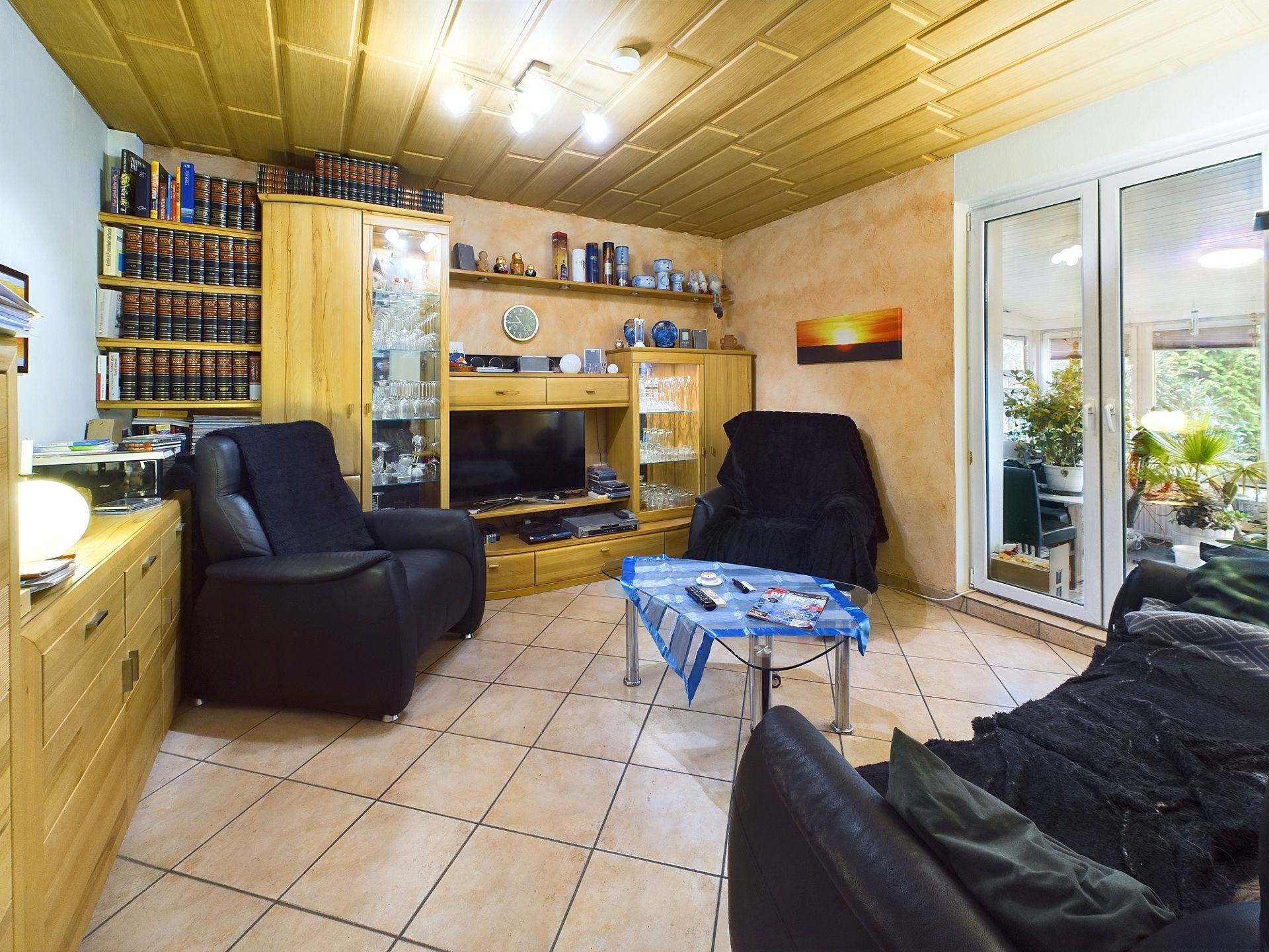 Wohnzimmer mit Ledersesseln, Holzmöbeln, Regalwand, Fernseher und Glastüren zum Wintergarten.