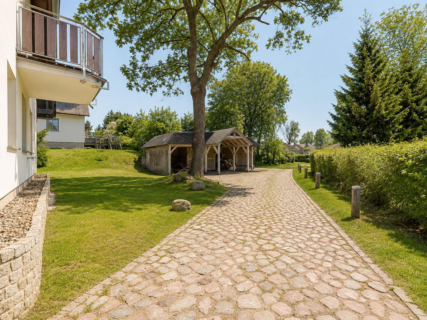 Pflasterweg mit Carport und Baum neben Wohnhaus, umgeben von Rasen und Büschen.