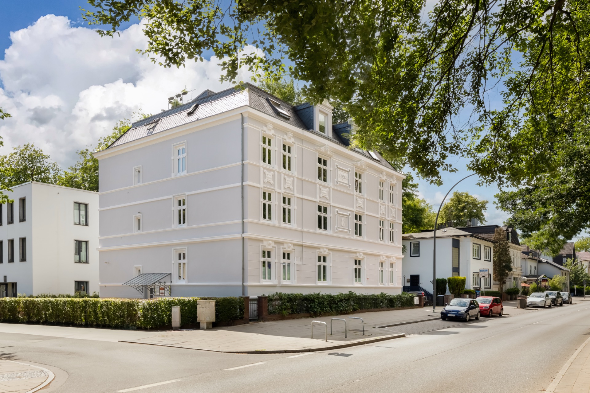 Weiße Wohnhausfassade mit vielen Fenstern an einer ruhigen Straße, im Vordergrund Bäume und parkende Autos.