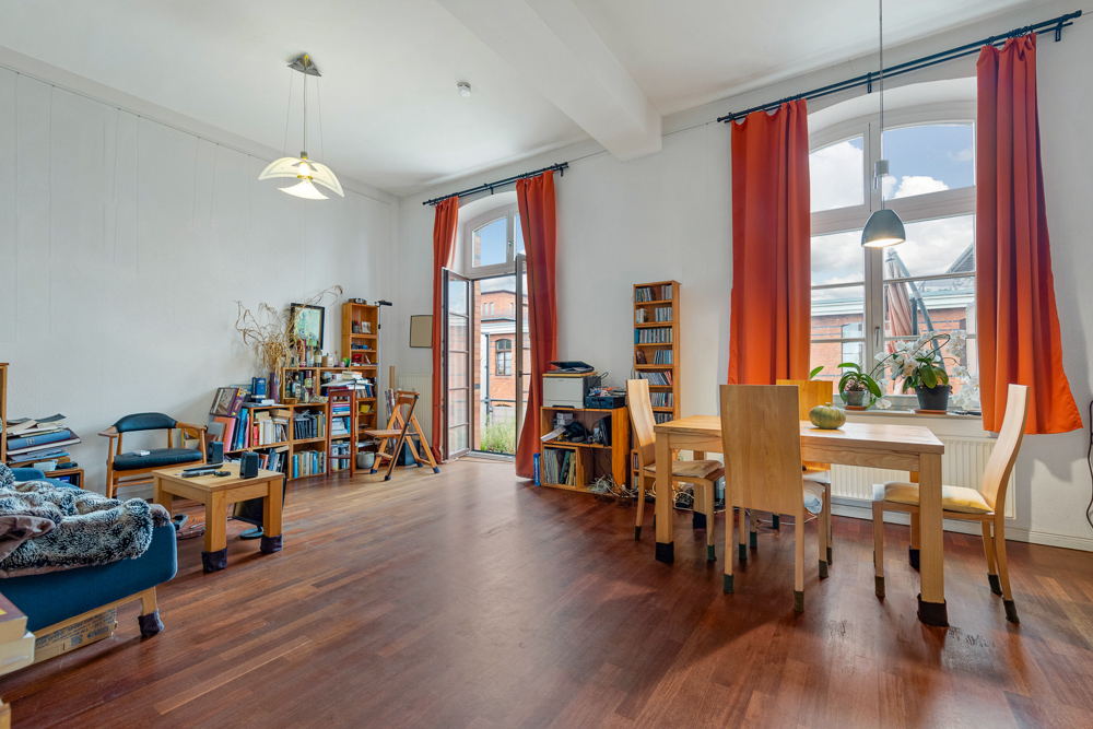 Wohnzimmer mit Holzboden, Esstisch, Bücherregalen und roten Vorhängen vor großen Fenstern.