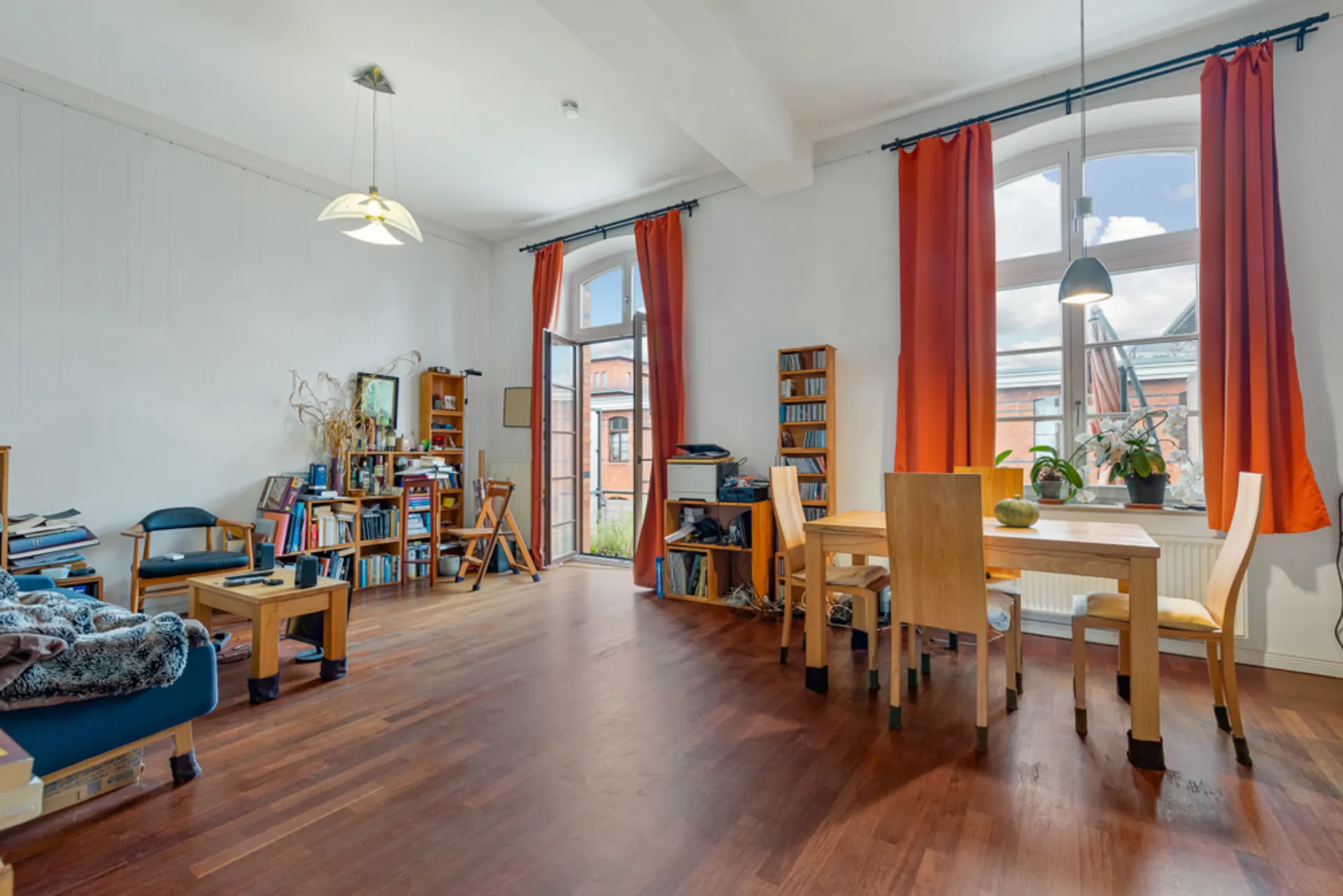 Wohnzimmer mit Holzboden, Esstisch, Bücherregalen und roten Vorhängen vor großen Fenstern.