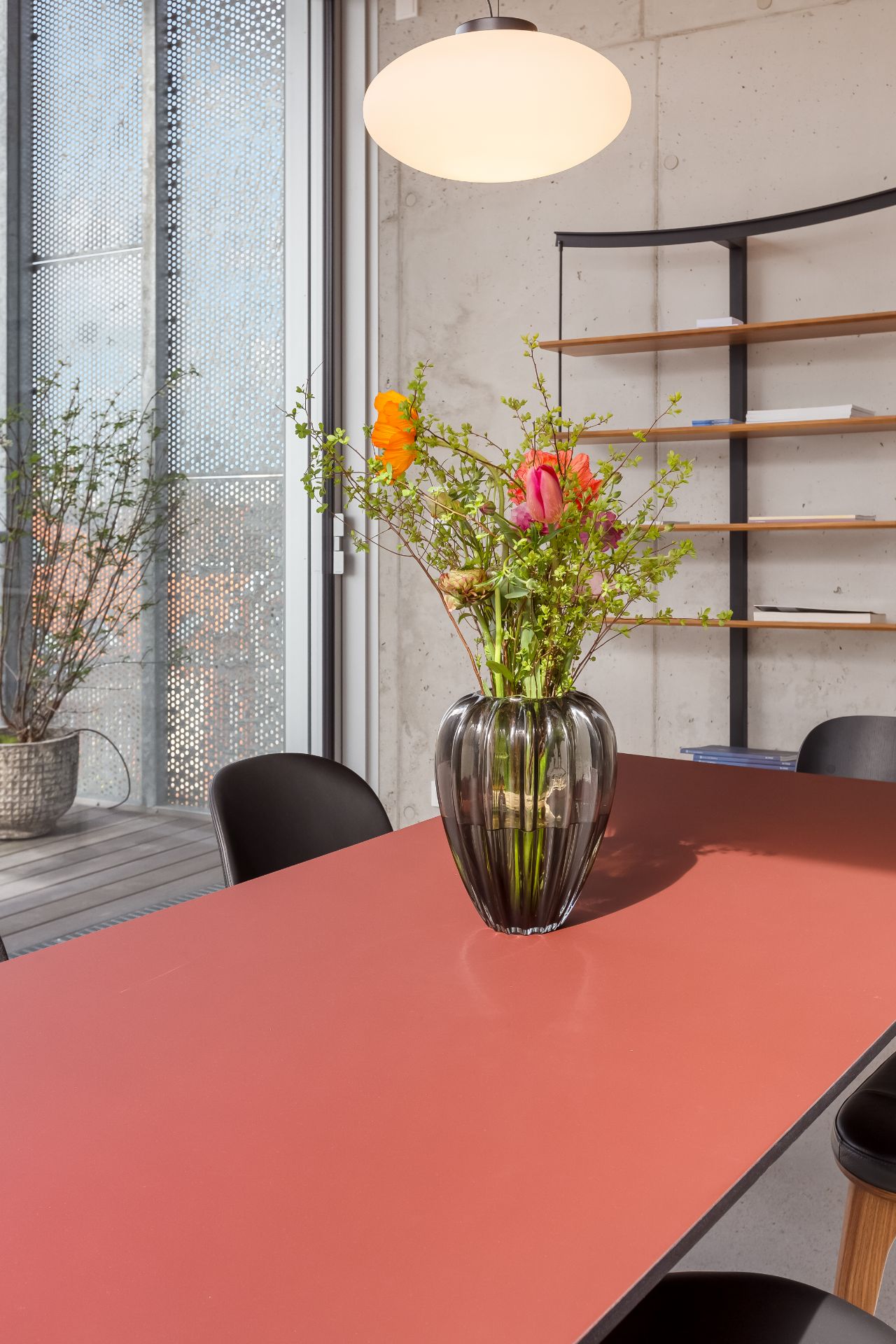 Rotbrauner Tisch mit Vase mit Blumen, schwarze Stühle, Regal und Lampe im Hintergrund.
