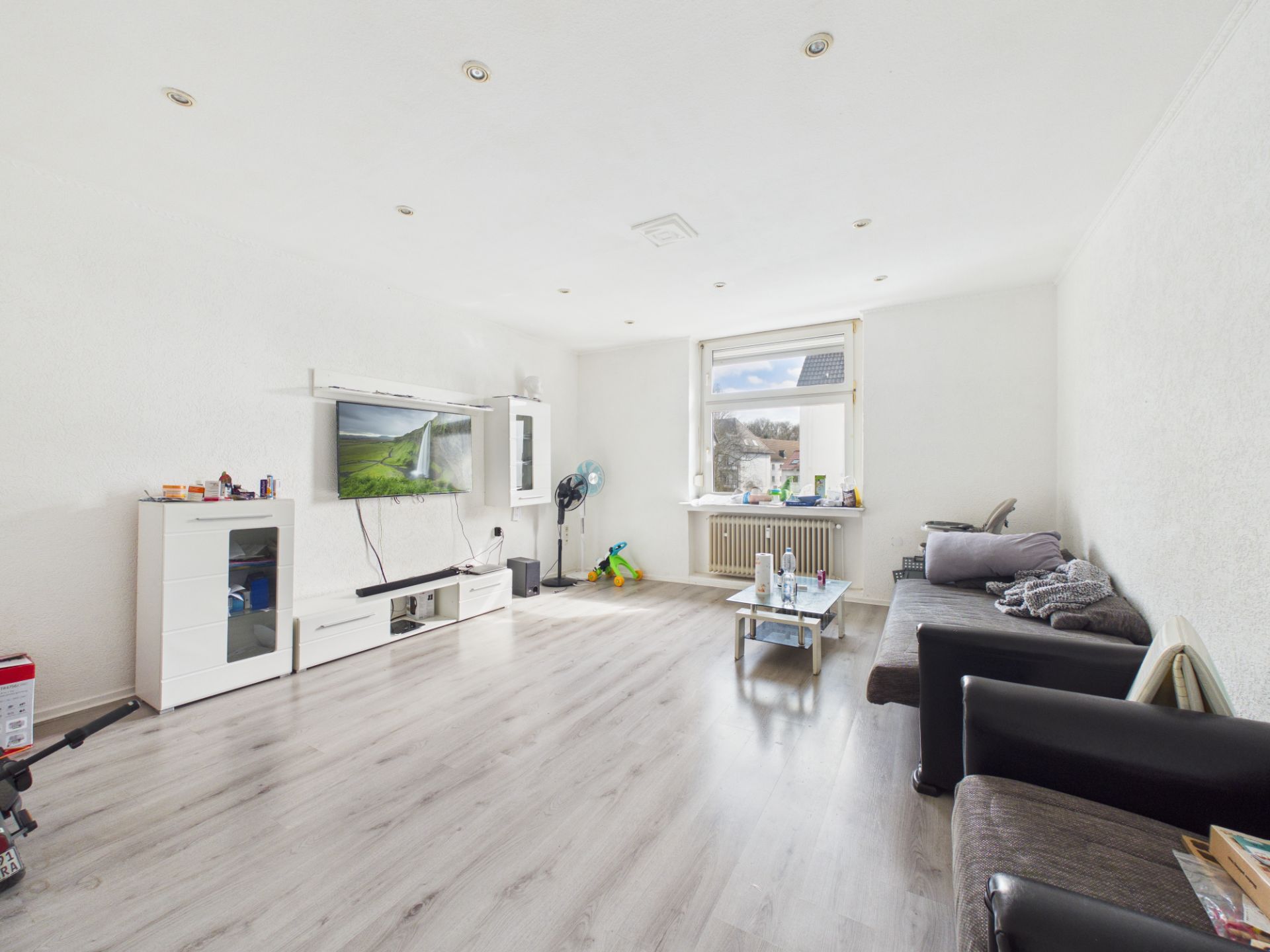 Helles Wohnzimmer mit Sofa, Fernseher, Glastisch und großem Fenster.