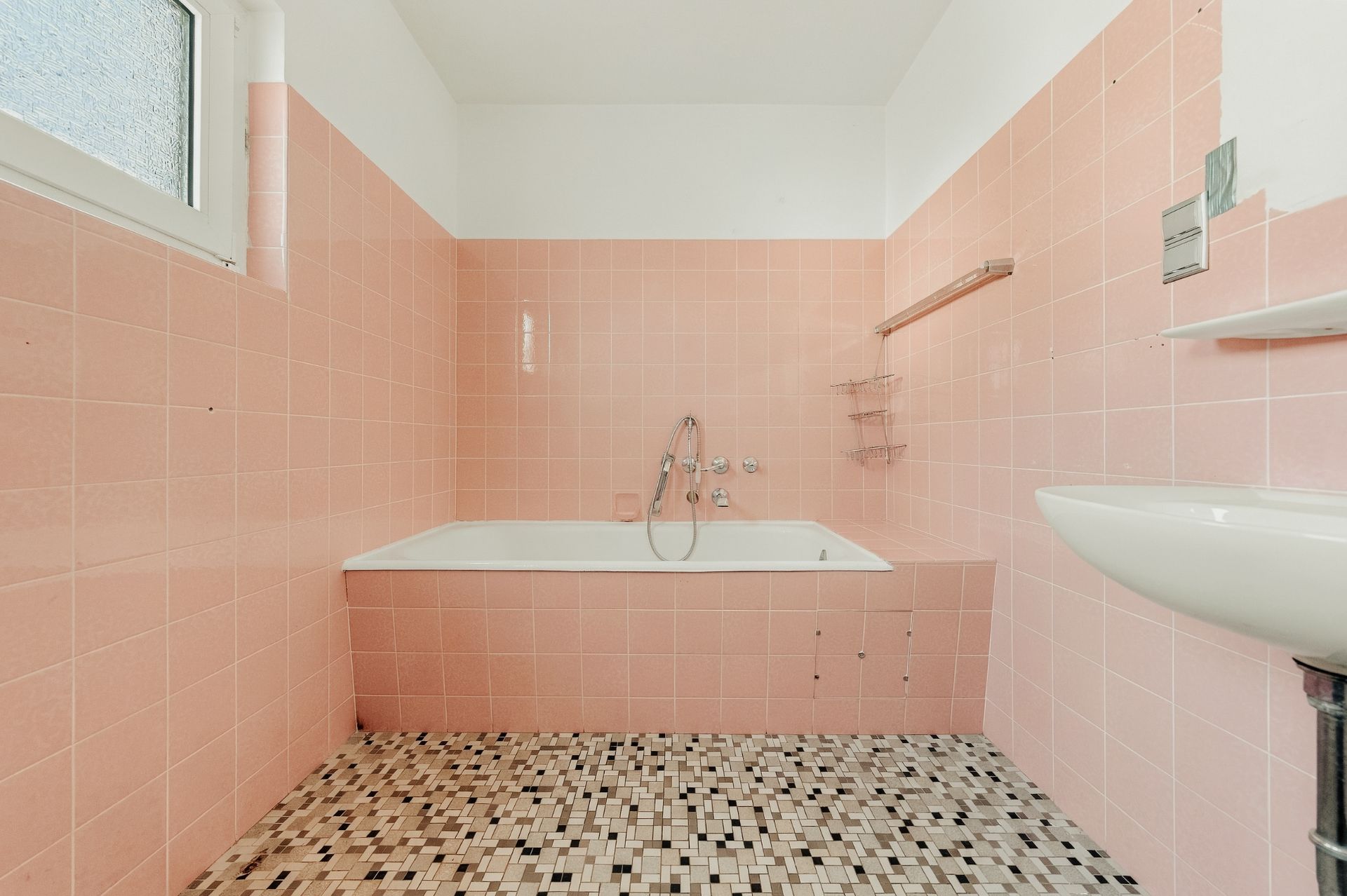 Badezimmer mit rosa Fliesen, Badewanne, kleinem Waschbecken und Fenster, Mosaikfliesen am Boden.