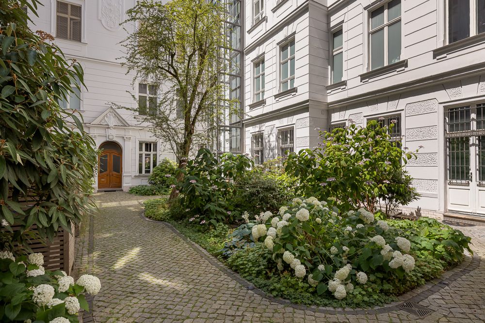 Gepflasterter Innenhof mit Blumenbeeten, Sträuchern und Bäumen vor einem Wohngebäude.