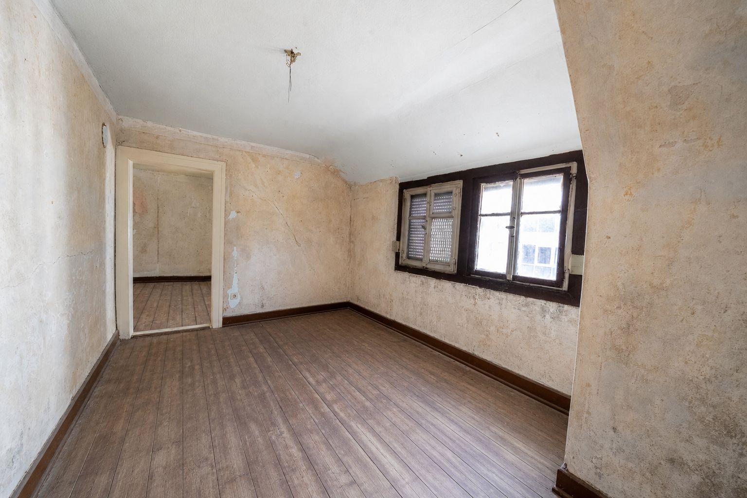 Leeres Zimmer mit Holzfußboden, abgenutzten Wänden und drei Fenstern, Tageslicht kommt herein.