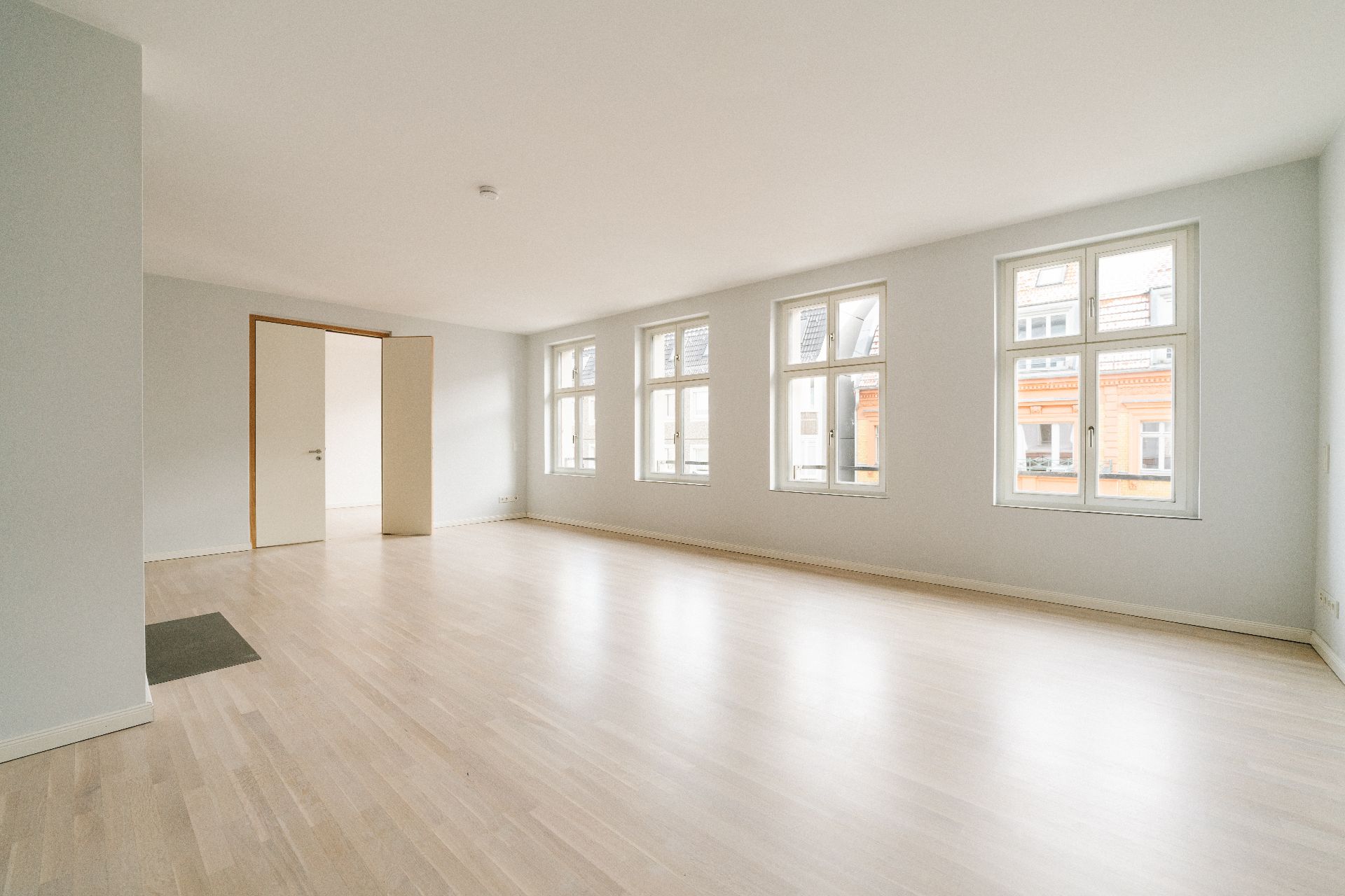Helles, leeres Zimmer mit vier großen Fenstern und Holzboden.