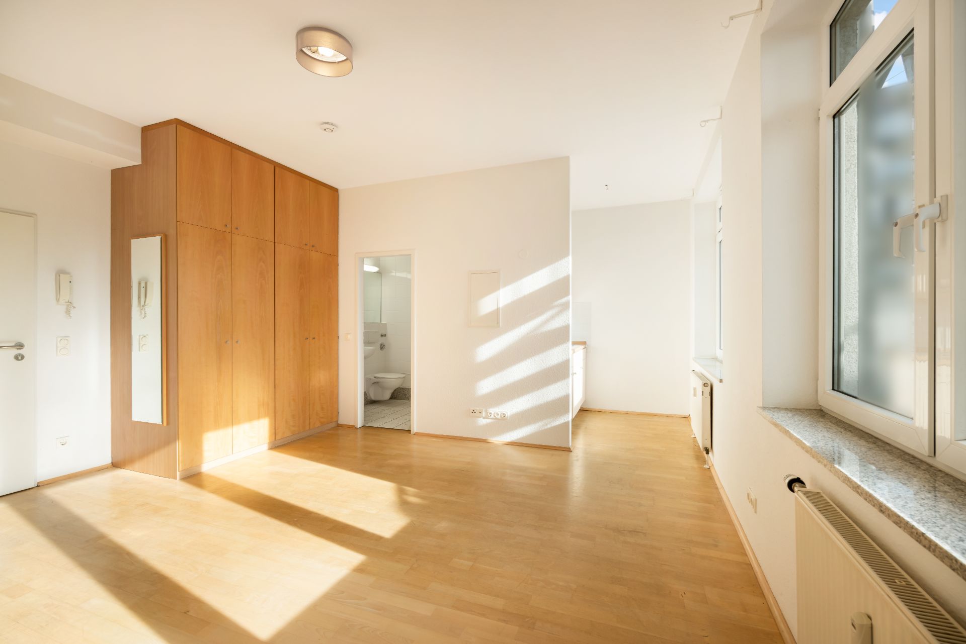 Heller Raum mit Holzboden, Einbauschrank, großen Fenstern und Blick ins Badezimmer.