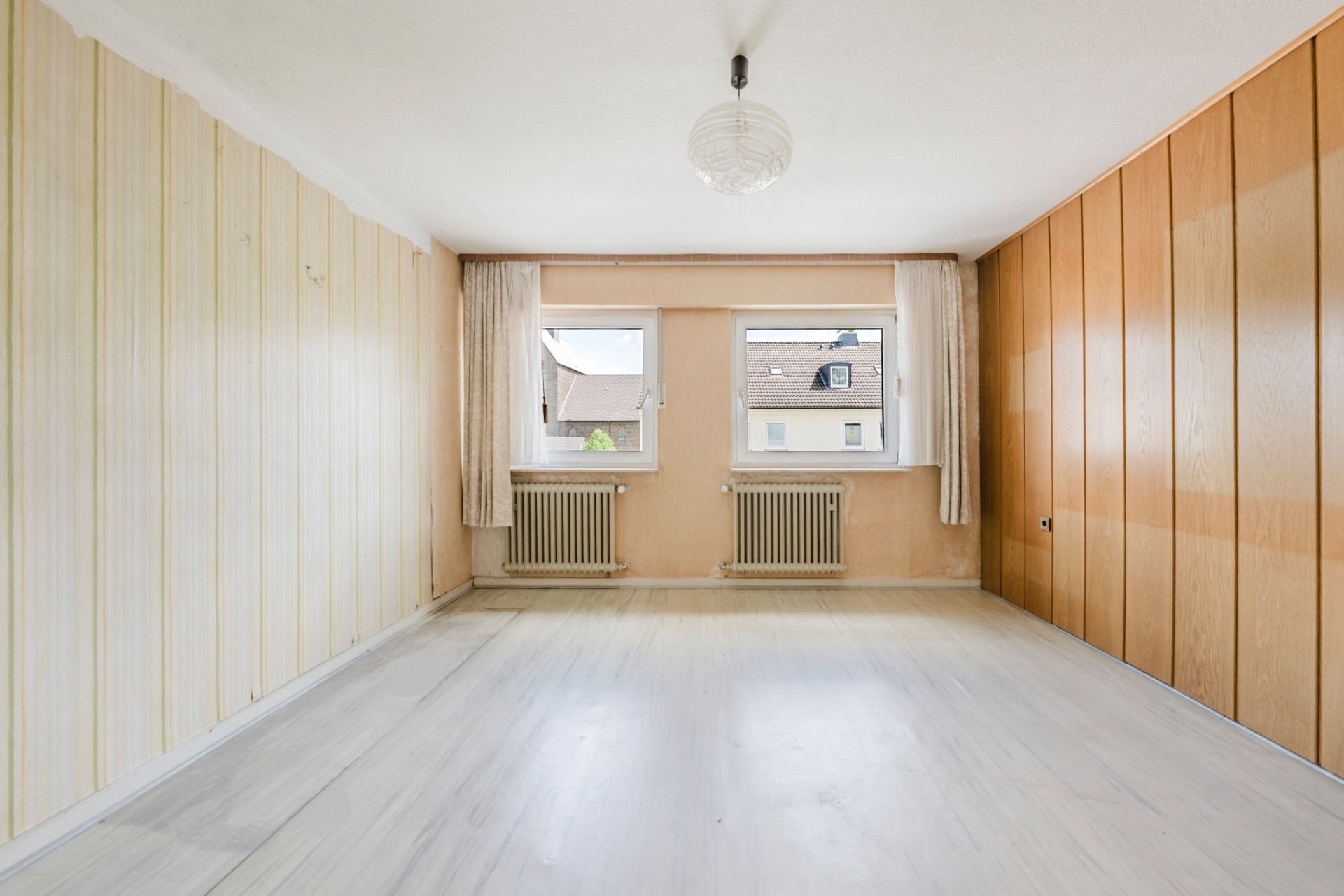 Leeres Zimmer mit zwei Fenstern, hellen Vorhängen, Radiatoren und Holzvertäfelung an einer Wand.