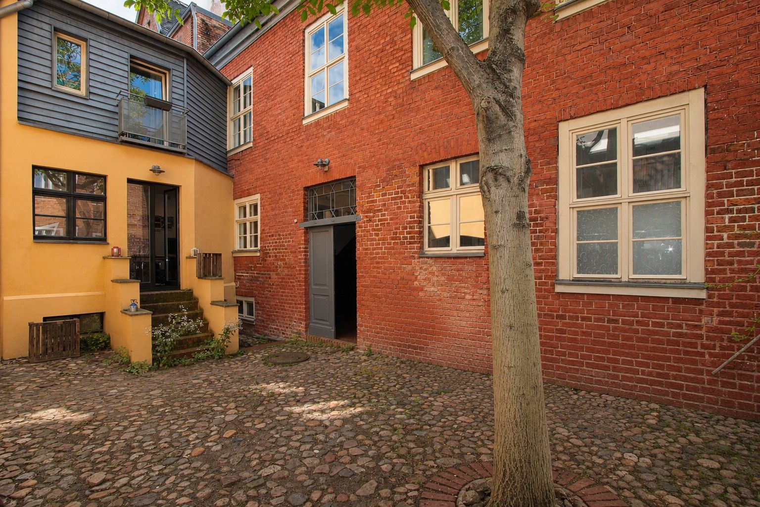 Gepflasterter Innenhof mit Baum vor roten und gelben Hausfassaden und mehreren Fenstern.