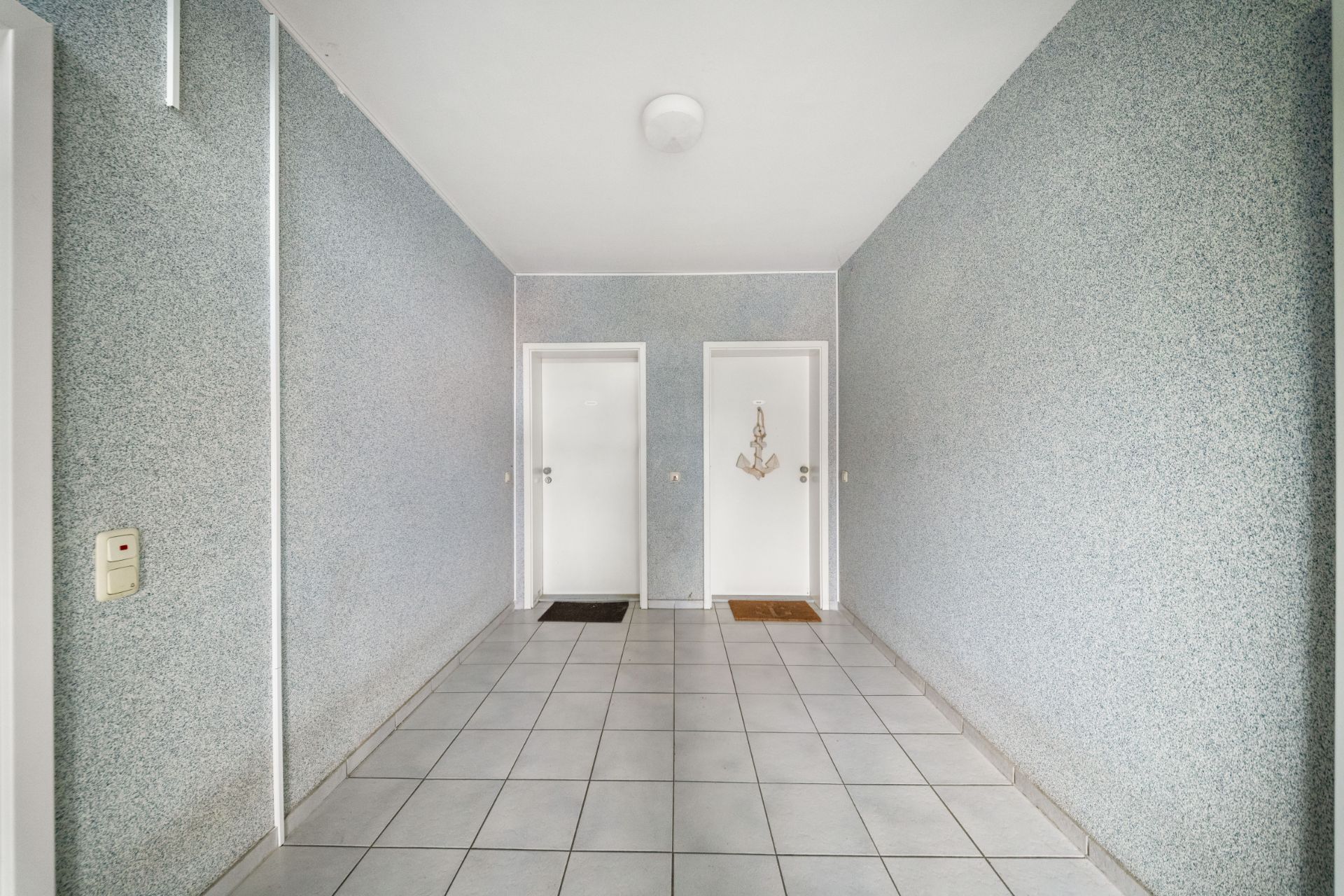 Flur mit zwei weißen Türen, grauen Wänden und Fliesenboden, jeweils mit Fußmatte davor.