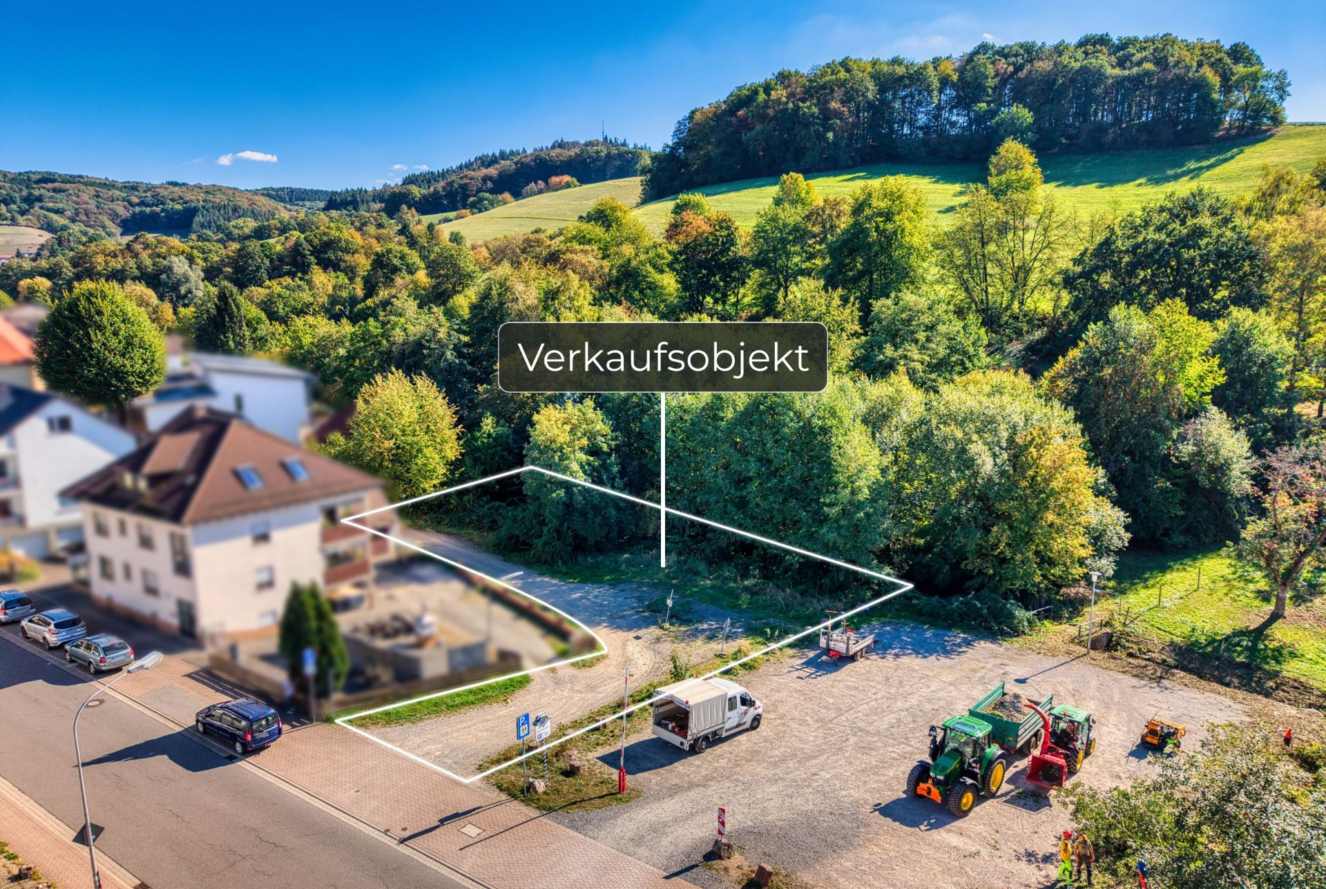 Leeres Grundstück mit weißer Markierung neben Wohnhäusern, Straßenfahrzeugen und Bäumen.