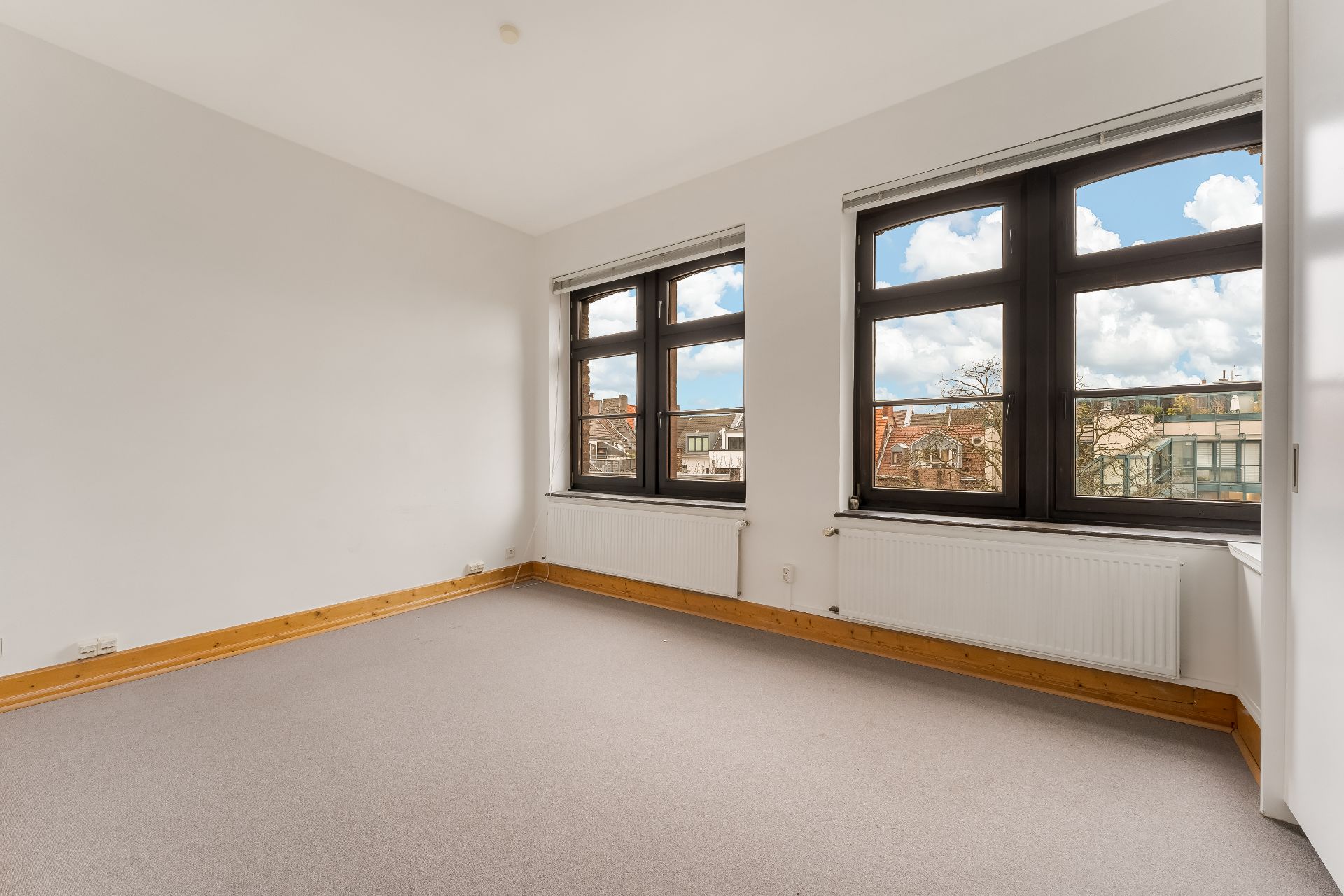 Leerer Raum mit zwei großen Fenstern, weiß gestrichenen Wänden und hellem Teppichboden.