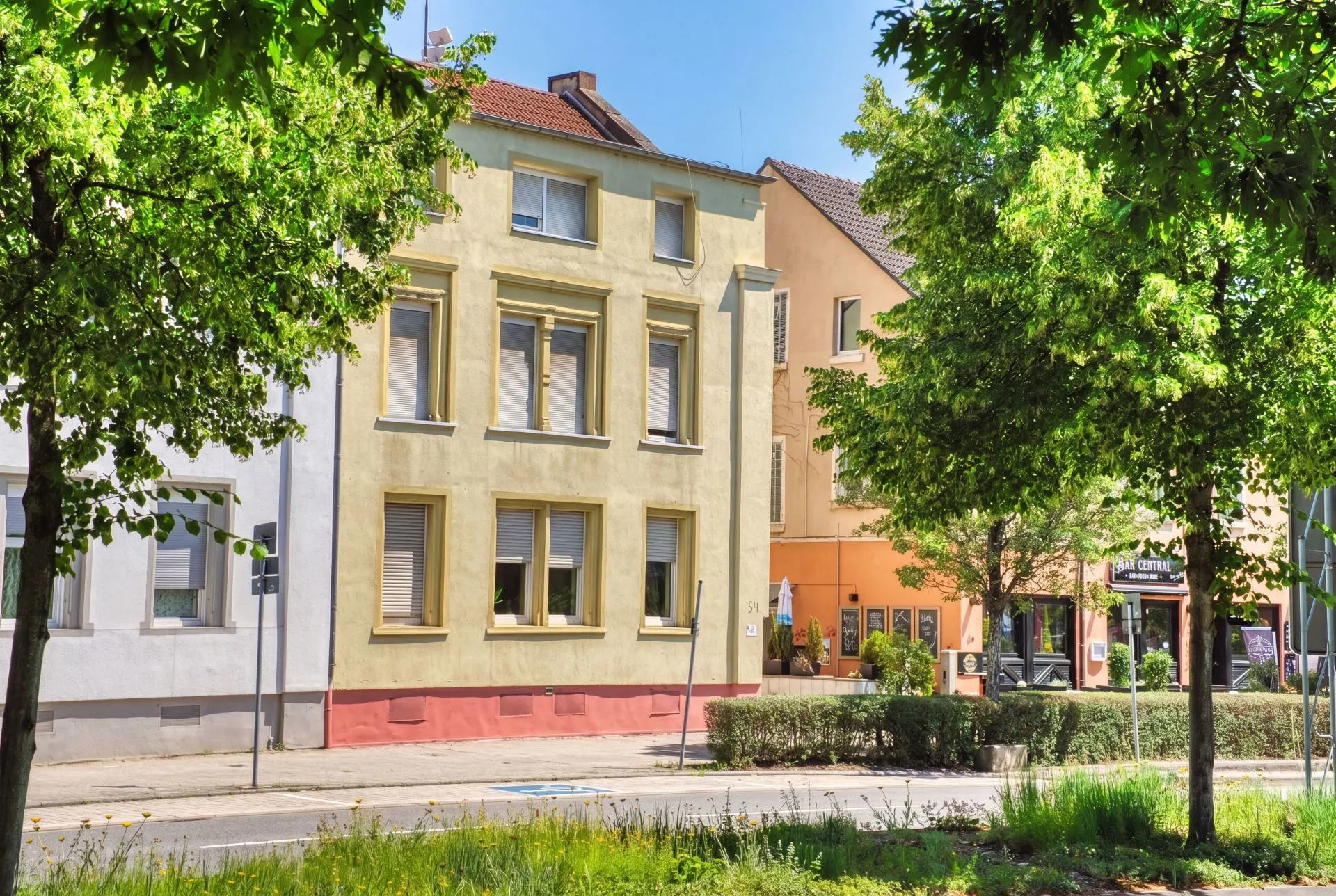 Mehrfamilienhaus mit gelber Fassade und mehreren Fenstern, vorne Bäume und Grünfläche.
