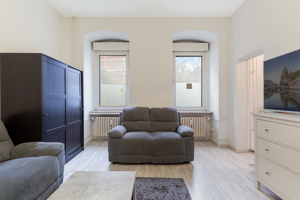 Wohnzimmer mit grauem Sofa, dunklem Schrank, TV auf Kommode und zwei großen Fenstern.