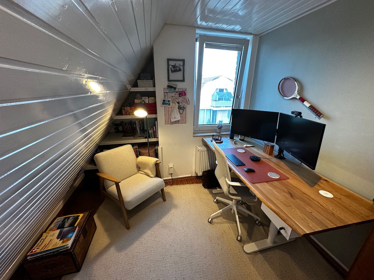 Kleines Arbeitszimmer mit Schreibtisch, zwei Monitoren, Sessel und Regal unter einer Dachschräge.
