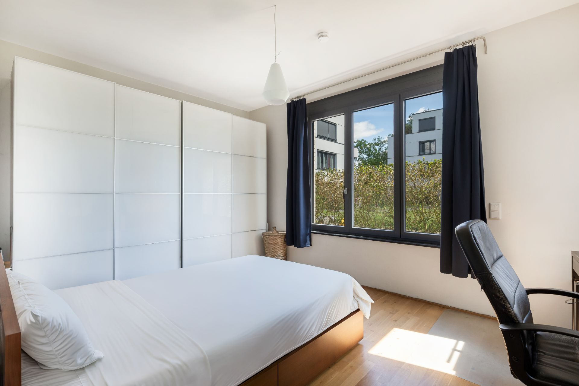 Helles Schlafzimmer mit großem Fenster, Doppelbett, weißem Kleiderschrank und Schreibtischstuhl.
