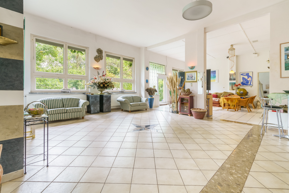 Helles Wohnzimmer mit Fliesenboden, zwei Sofas, großen Fenstern und Essbereich im Hintergrund.
