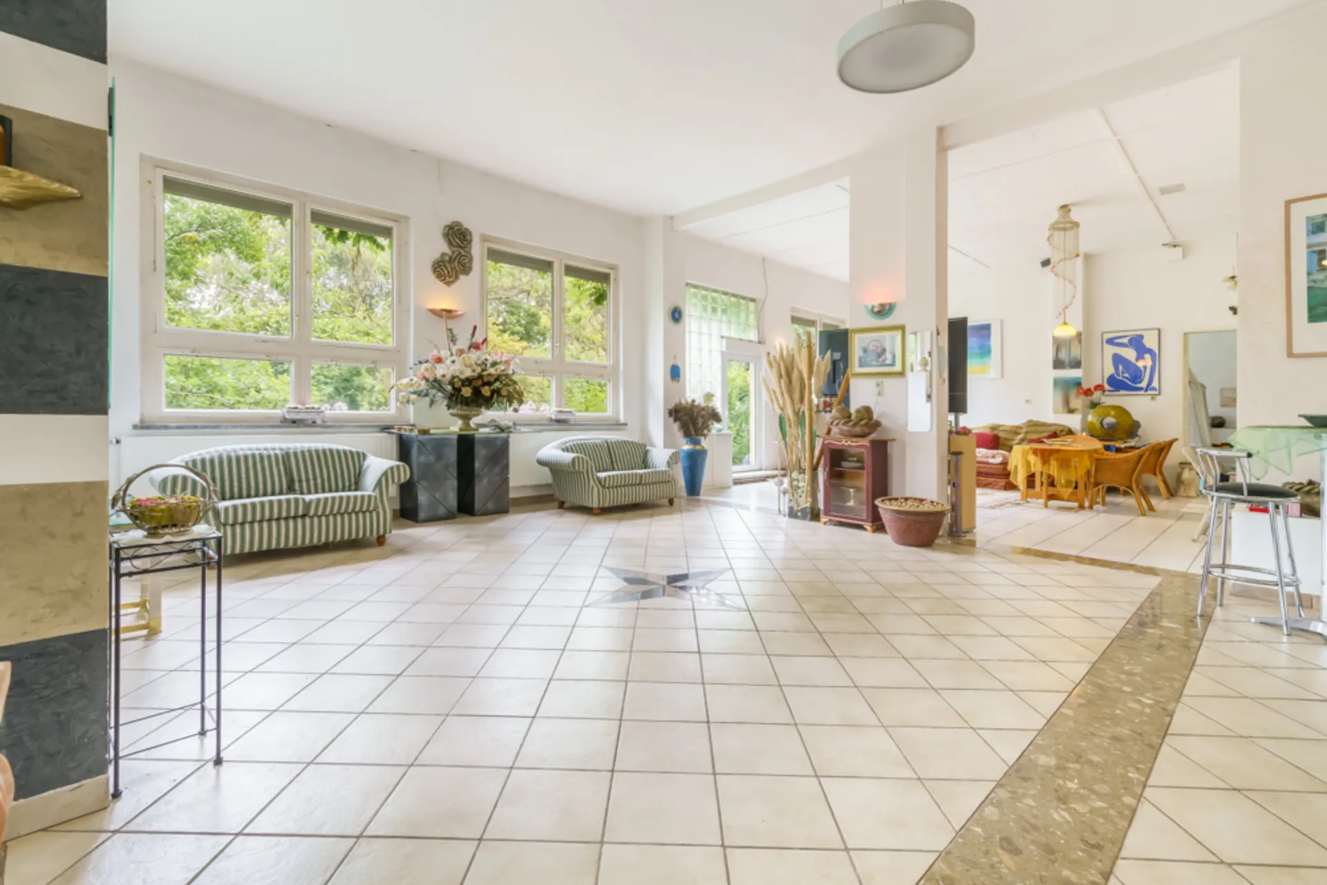 Helles Wohnzimmer mit Fliesenboden, zwei Sofas, großen Fenstern und Essbereich im Hintergrund.